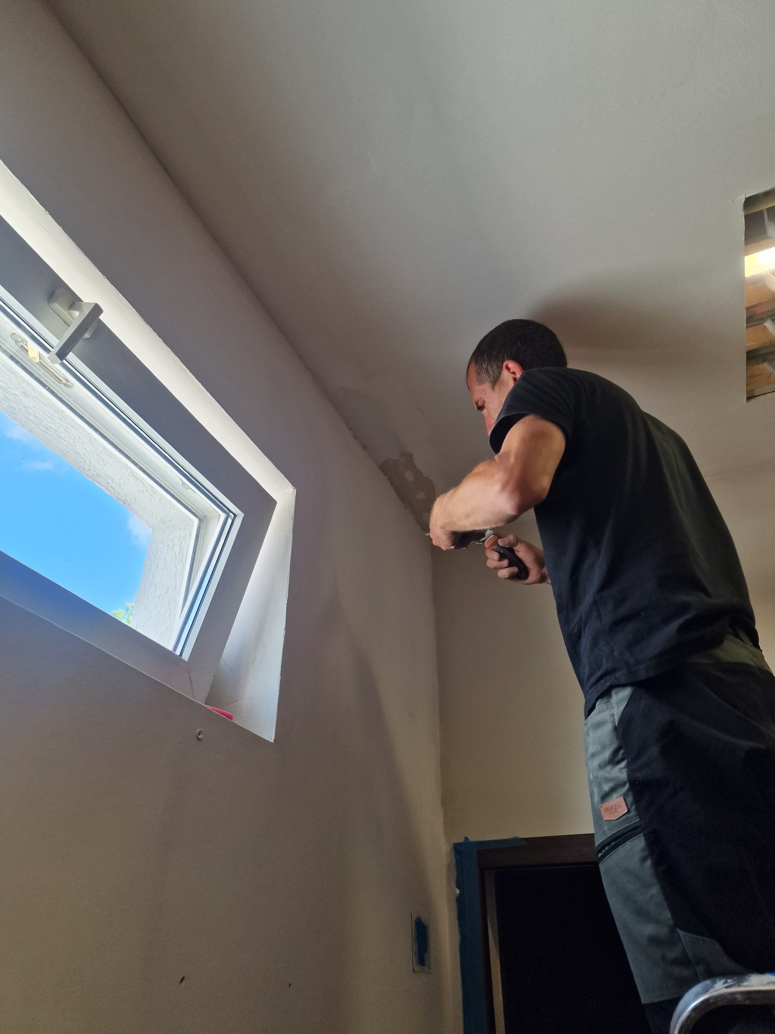 Mężczyzna na drabinie szpachluje fragment ściany pod oknem dachowym, widoczne niedoskonałości i nierówności na powierzchni przed malowaniem.