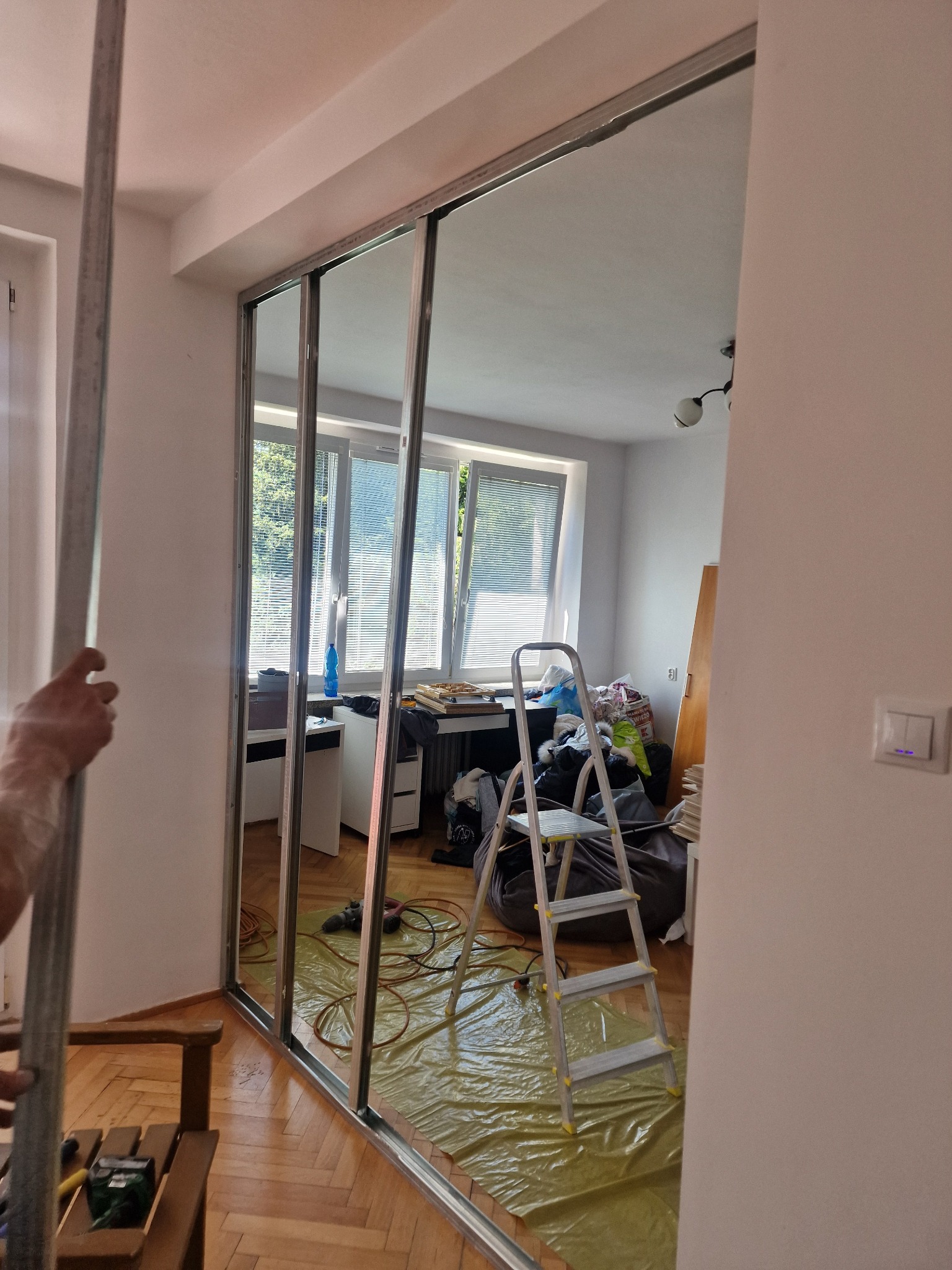 Montaż ramy przesuwnej szafy wnękowej z lustrami, widoczne narzędzia i zabezpieczona folią podłoga w pokoju z bałaganem.