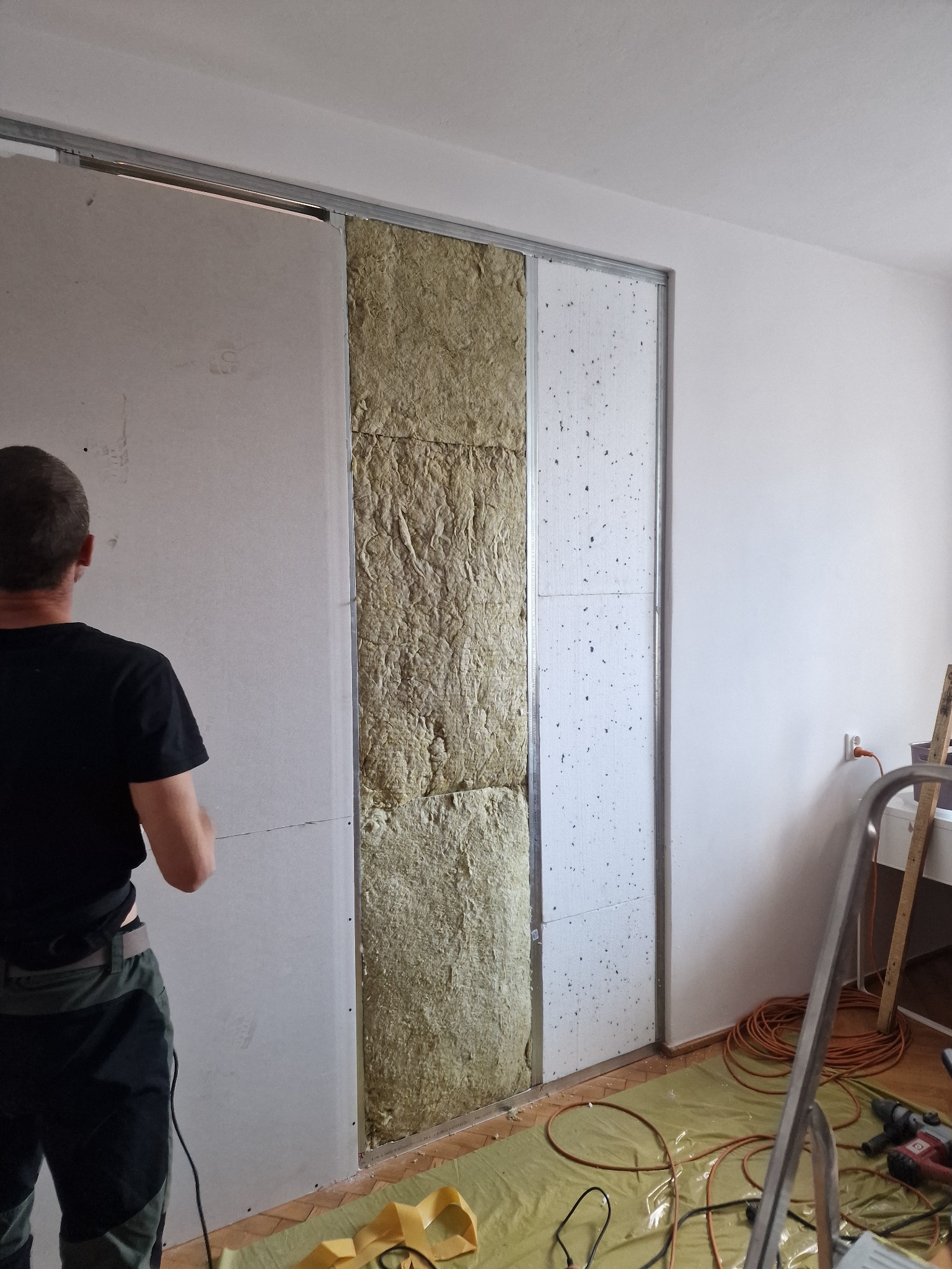 Prace wykończeniowe: montaż płyty gipsowo-kartonowej na ścianie z ociepleniem z wełny mineralnej, widoczne profile metalowe, narzędzia i zabezpieczenia podłogi.