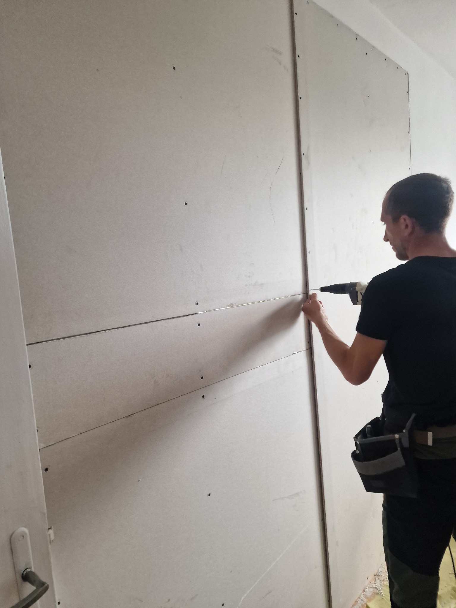 Mężczyzna w czarnej koszulce przykręca płytę gipsowo-kartonową do ściany za pomocą wkrętarki, widoczny pas z narzędziami, fragment drzwi po lewej stronie.