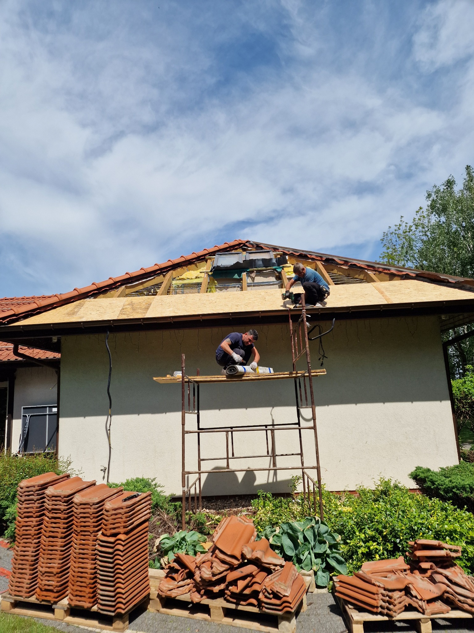 Dwóch dekarzy na rusztowaniu montuje nowe poszycie dachu z płyt OSB, widoczne stosy dachówek ceramicznych na paletach przed budynkiem.