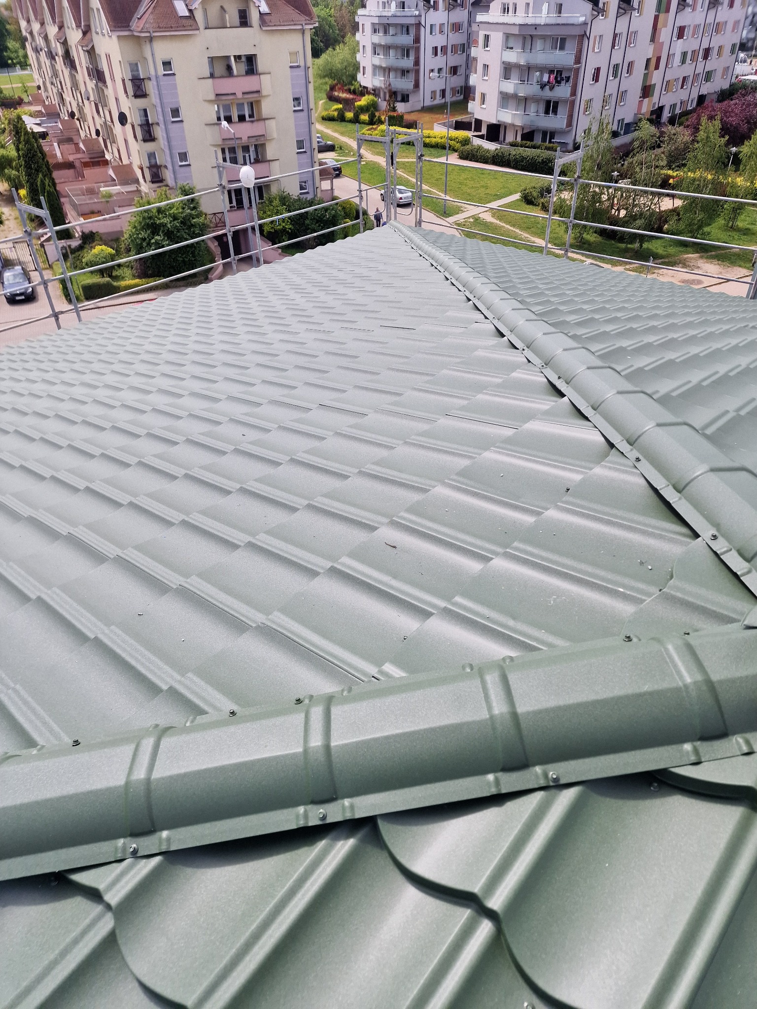 Nowy, zielony dach z blachodachówki modułowej na tle budynków mieszkalnych i rusztowania zabezpieczającego krawędź.