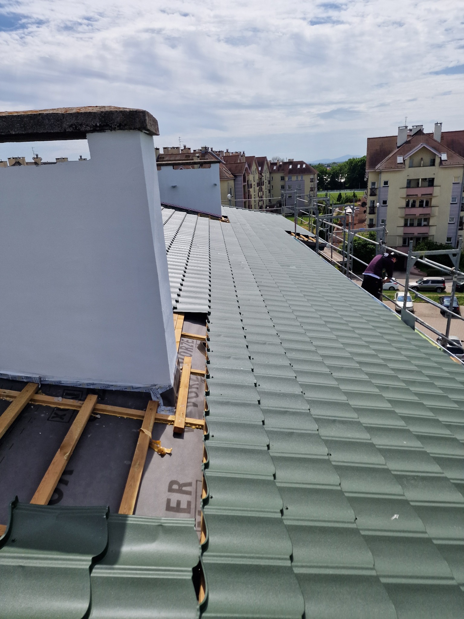 Montaż zielonej blachodachówki na dachu domu wielorodzinnego, widoczne rusztowanie, ocieplony komin i elementy drewniane konstrukcji dachu.