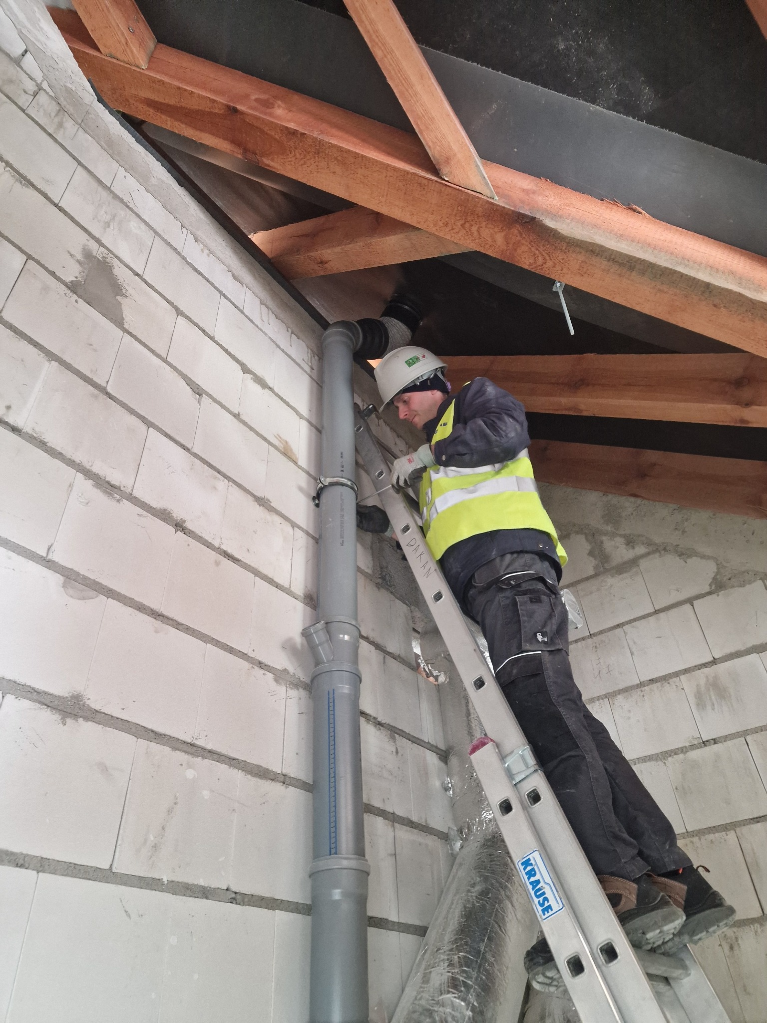 Pracownik w kasku i kamizelce odblaskowej na drabinie montuje pion kanalizacyjny w surowym wnętrzu poddasza z widoczną konstrukcją dachu i ścianami z betonu komórkowego.