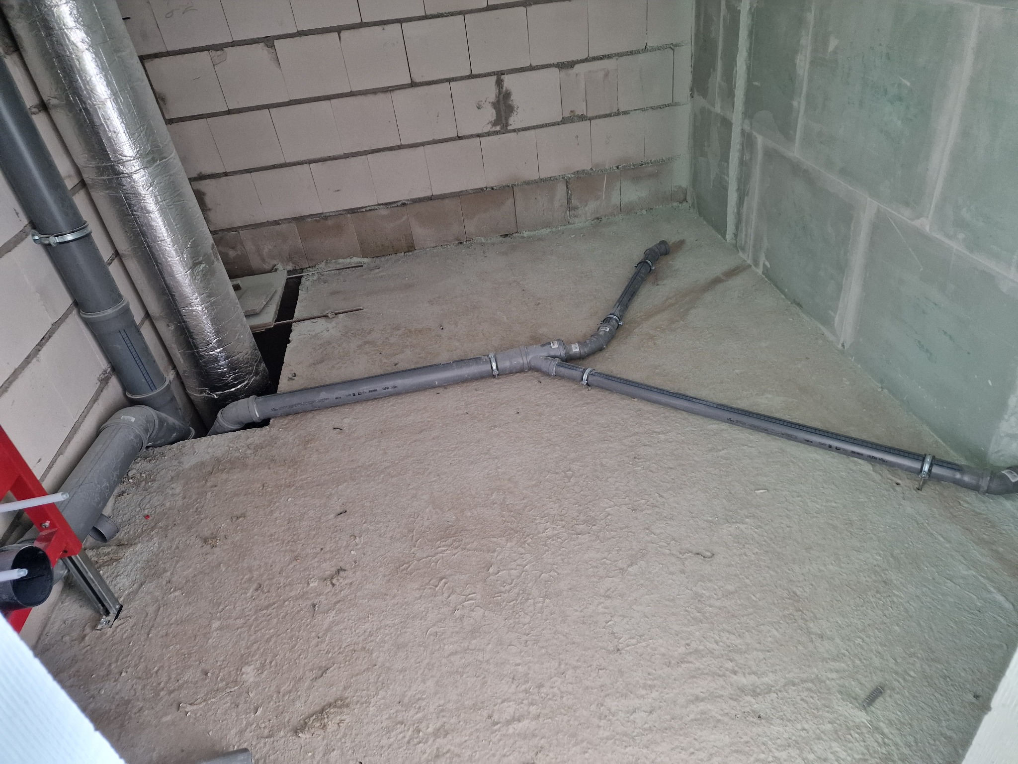 Wnętrze w stanie surowym z widocznymi rurami kanalizacyjnymi z szarego PCV, fragmentem drabiny oraz ocieplonym przewodem wentylacyjnym, ściany z betonu komórkowego i płyt gipsowo-kartonowych.