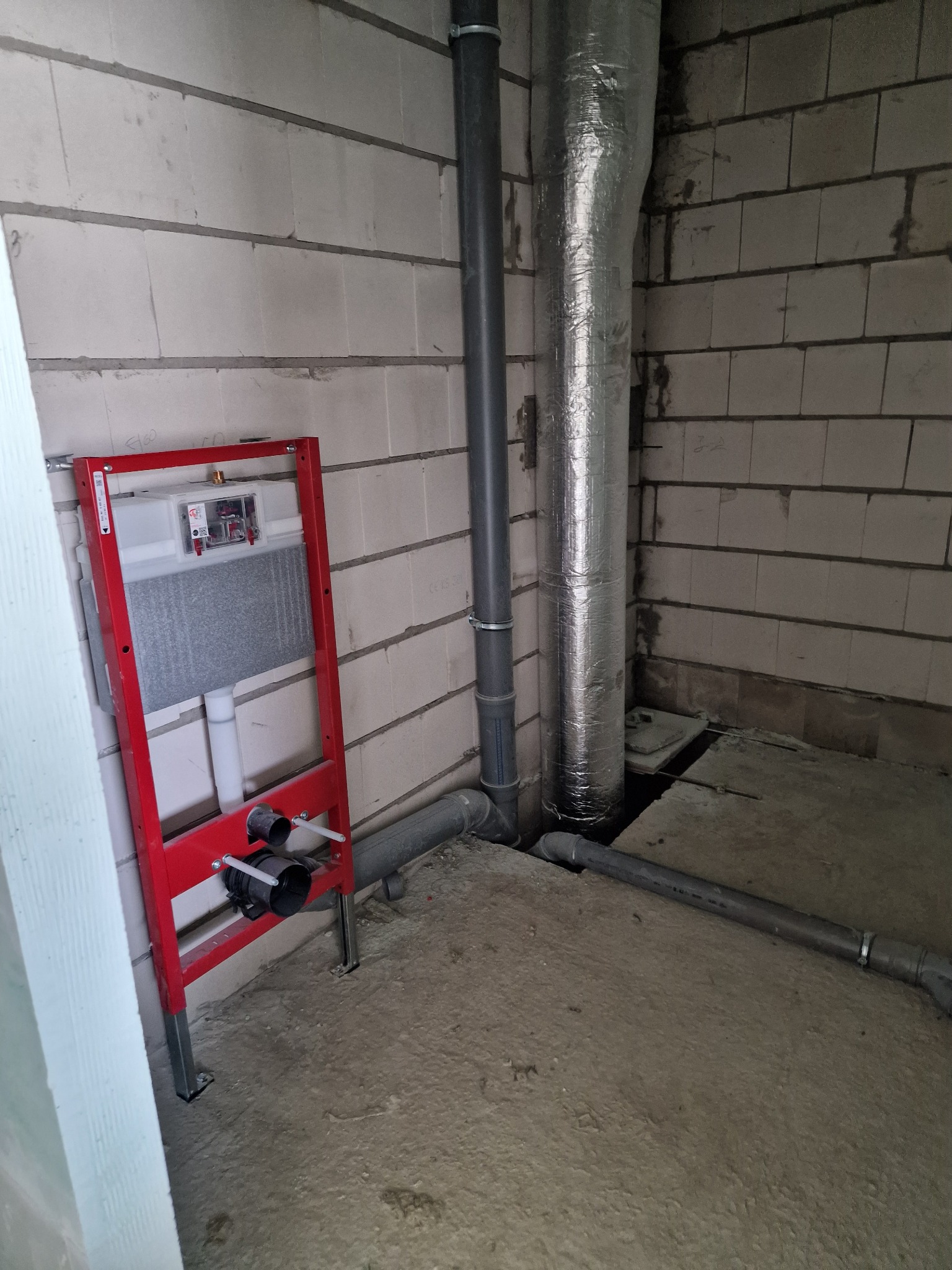 Stelaż podtynkowy WC z czerwonym profilem zamontowany w surowym pomieszczeniu z instalacjami wentylacyjnymi i kanalizacyjnymi na tle ściany z betonu komórkowego.