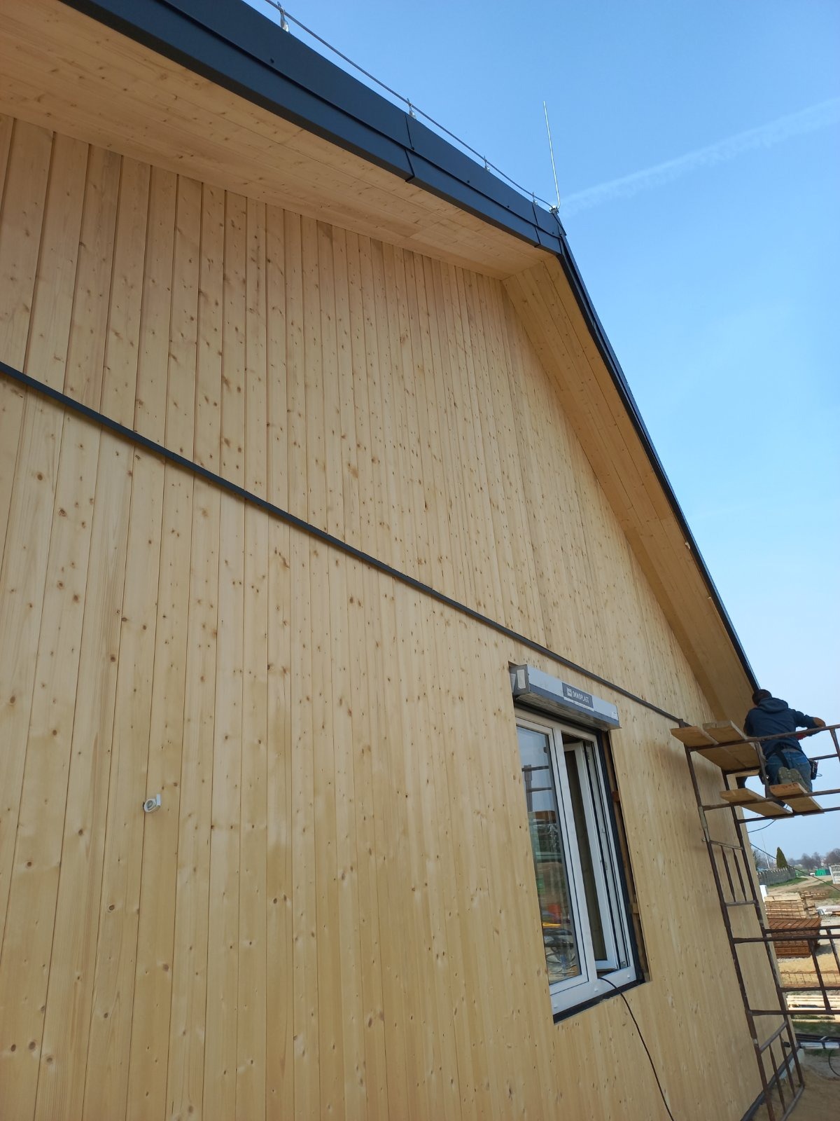 Montaż drewnianej elewacji na budynku jednorodzinnym, widoczny pracownik na rusztowaniu, okno z roletą zewnętrzną i elementy dachu.