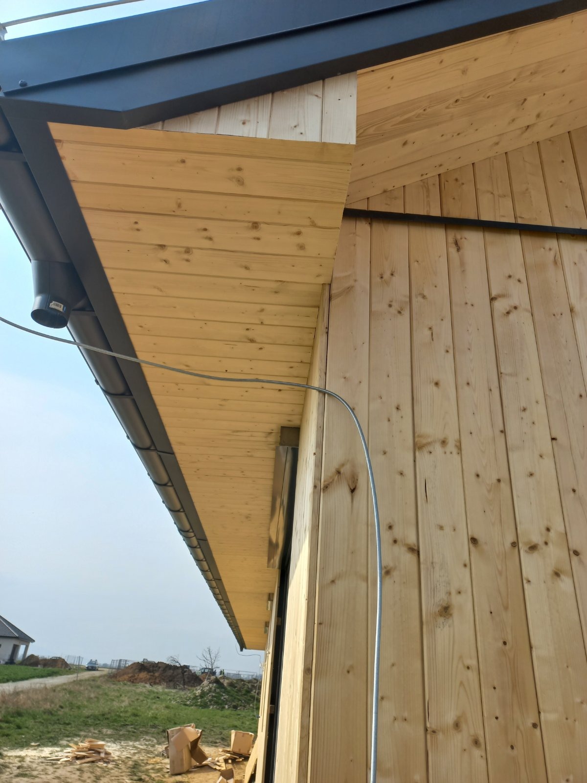 Drewniana elewacja budynku w trakcie budowy z widocznymi elementami dachu i rynną, na tle krajobrazu z domem i stertą ziemi.