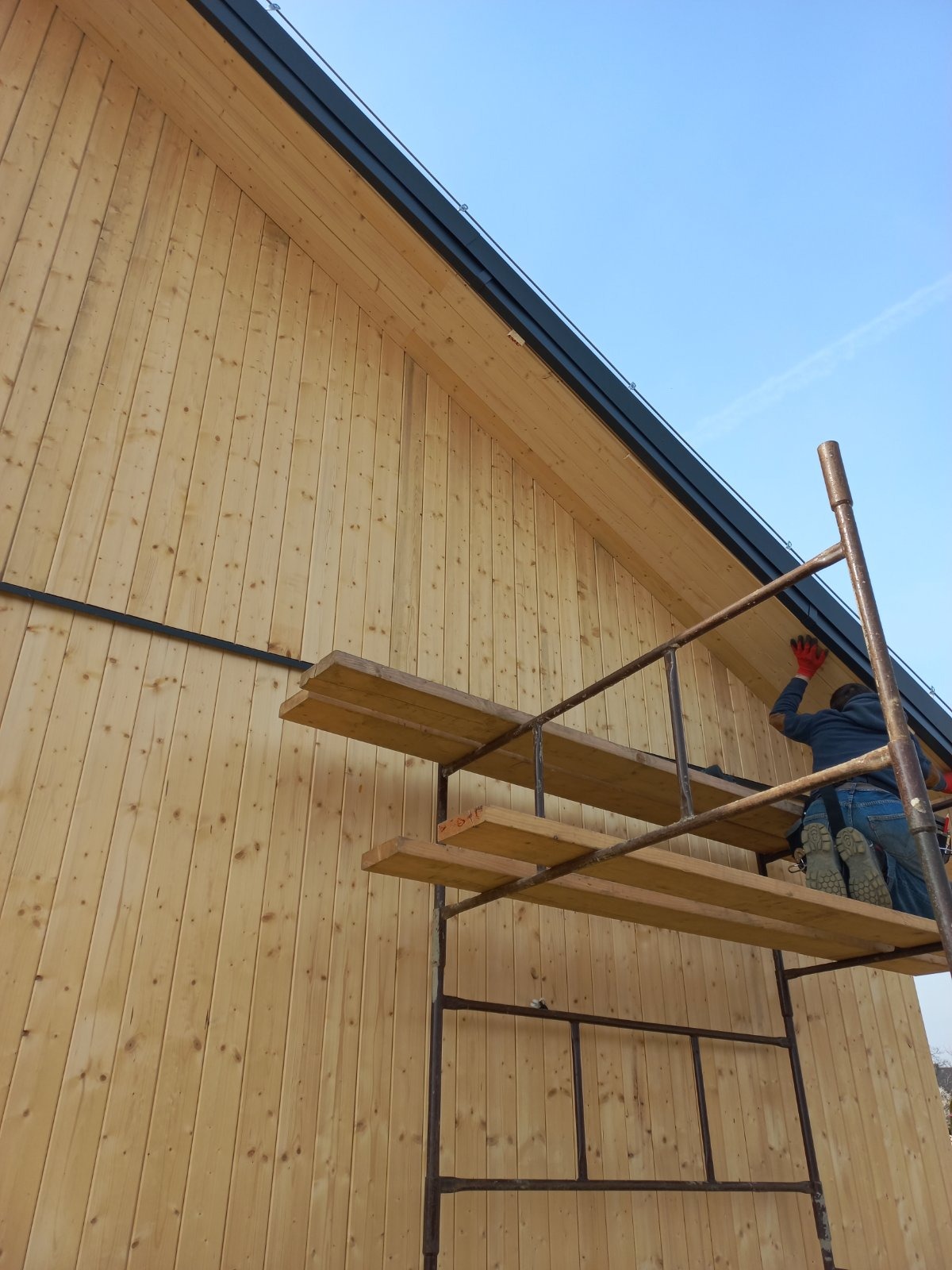 Pracownik w Gniechowicach na rusztowaniu montuje drewnianą elewację pionową z widoczną rynną dachową i obróbką blacharską.