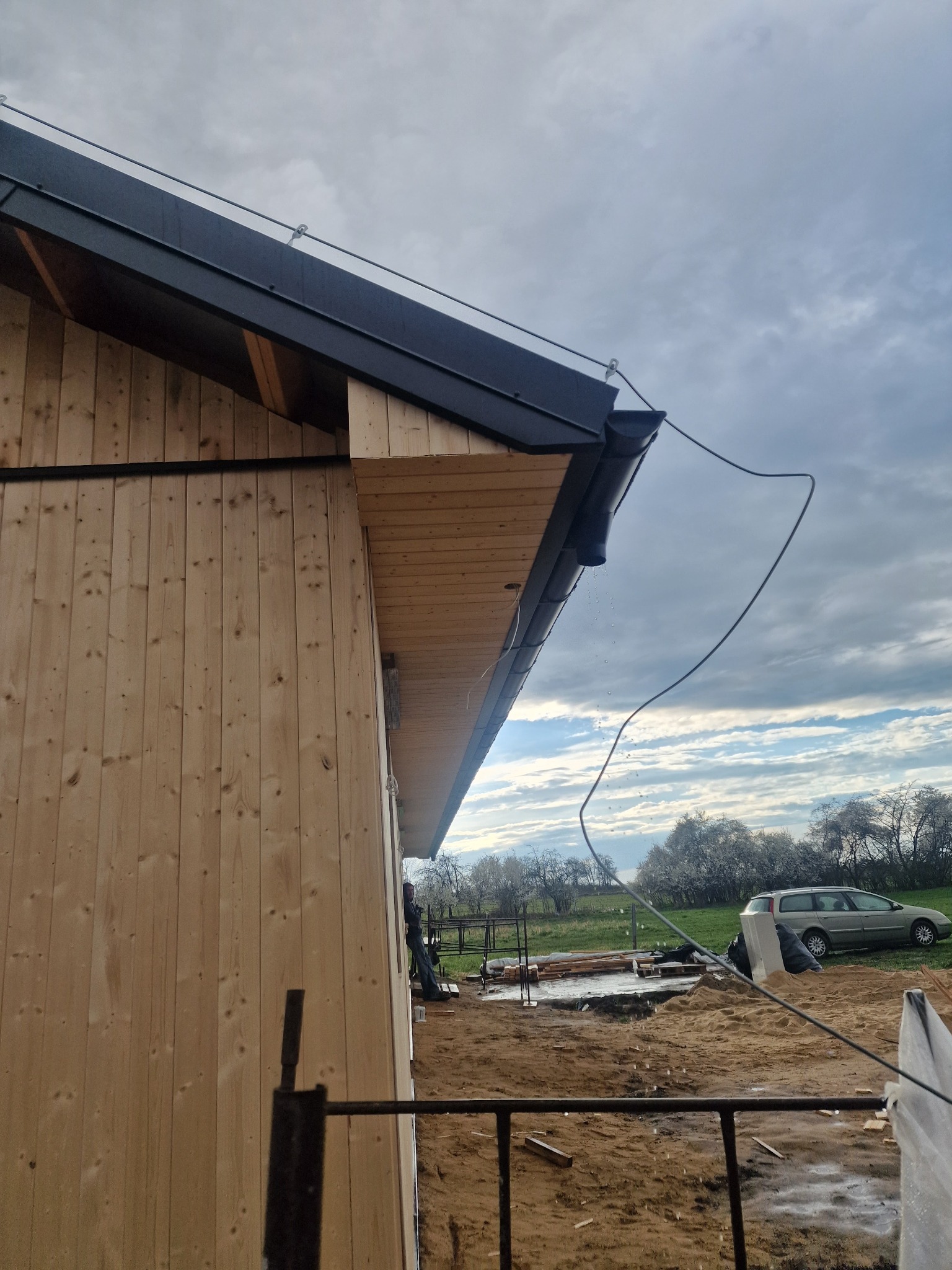 Nowe drewniane poszycie domu z czarnym dachem i rynnami w trakcie montażu, widoczne krople wody z rynny i kabel zwisający z dachu, w tle zachmurzone niebo i samochód.
