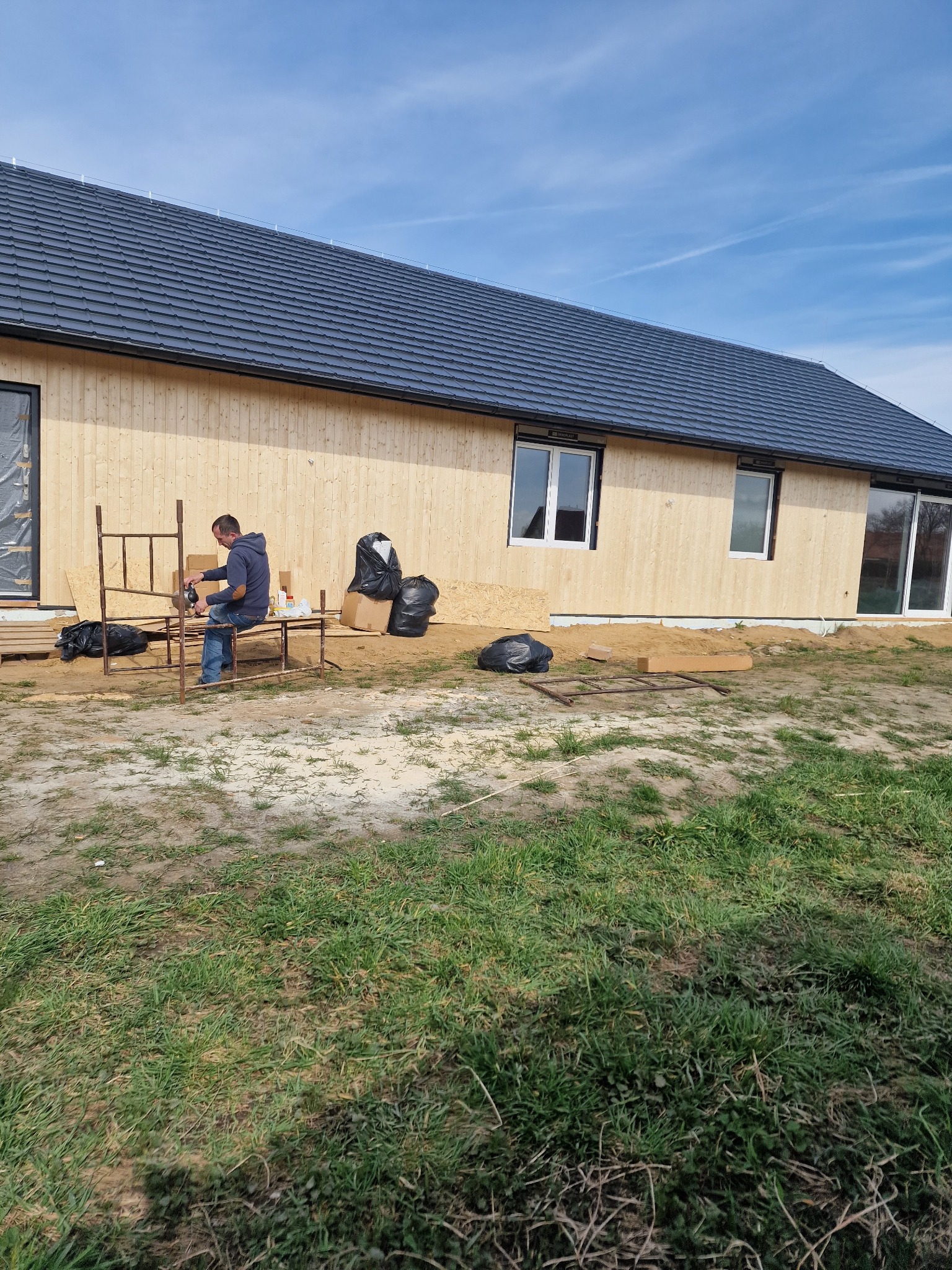 Pracownik w bluzie i jeansach, na prowizorycznym rusztowaniu, obrabia element blacharski na tle domu z ciemnym dachem i jasną elewacją z desek.