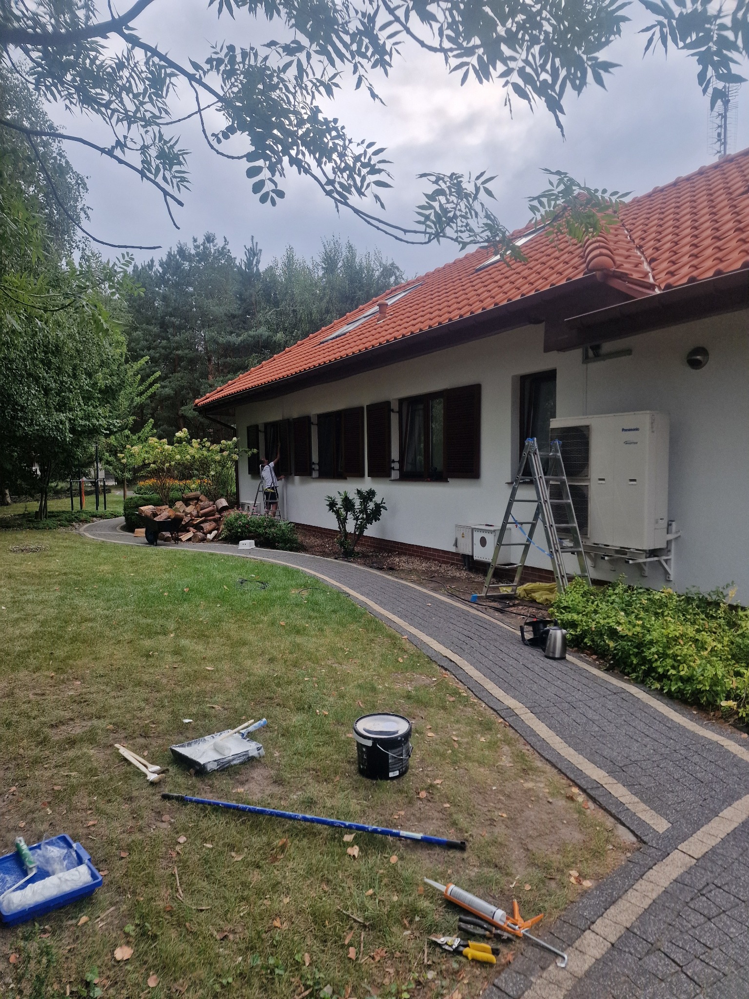 Malowanie elewacji domu jednorodzinnego z brązowymi okiennicami, widoczne narzędzia malarskie rozłożone na trawniku, drabina oparta o ścianę, w tle wózek z drewnem.