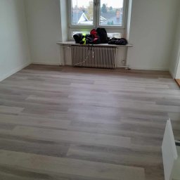 Dominik Olesiak - Jasnoszara podłoga z paneli ułożona w pustym pokoju z białymi ścianami, grzejnikiem pod oknem i torbami na parapecie.