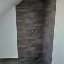 Dominik Olesiak - Ukończona kabina prysznicowa w łazience na poddaszu, wyłożona szarymi płytkami imitującymi kamień z brązowymi akcentami, widoczny odpływ liniowy i przygotowane przyłącza wodne.