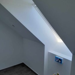 Dominik Olesiak - Poddasze po odświeżeniu: białe ściany i sufit, okno dachowe w skosie, szare płytki podłogowe, częściowo zamaskowana instalacja sanitarna na ścianie.