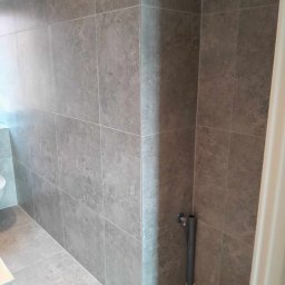 Dominik Olesiak - Szare, matowe płytki ceramiczne wielkoformatowe na ścianach i podłodze w rogu łazienki, widoczna rura instalacyjna przy ścianie.