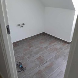 Dominik Olesiak - Płytki podłogowe w odcieniach szarości i brązu w małym pomieszczeniu, widoczne przyłącza wodne na ścianie, kabel elektryczny leżący na podłodze, widok zza uchylonych drzwi.