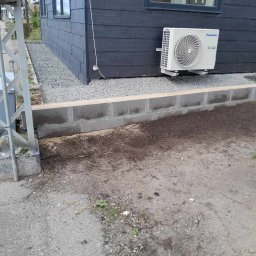 Dominik Olesiak - Ciemna elewacja domu z zamontowanym klimatyzatorem Panasonic, obok widoczny fundament z betonowych bloczków i nasypany żwir.