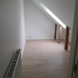 Dominik Olesiak - Poddasze po remoncie w Sławnie: świeżo pomalowane ściany na biało, jasne panele podłogowe, odsłonięte drewniane belki konstrukcyjne i grzejnik przy ścianie.