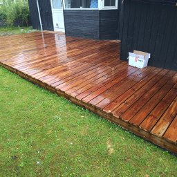 Dominik Olesiak - Świeżo zaimpregnowany, mokry drewniany taras przylegający do czarnej ściany budynku, obok kartonowe pudełko z napisem NKT.