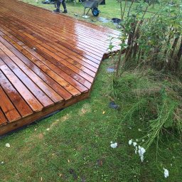 Dominik Olesiak - Świeżo zaimpregnowany drewniany taras z widoczną strukturą drewna, graniczący z trawnikiem i ozdobnym płotkiem z gałęzi, z pracownikiem i taczką w tle.