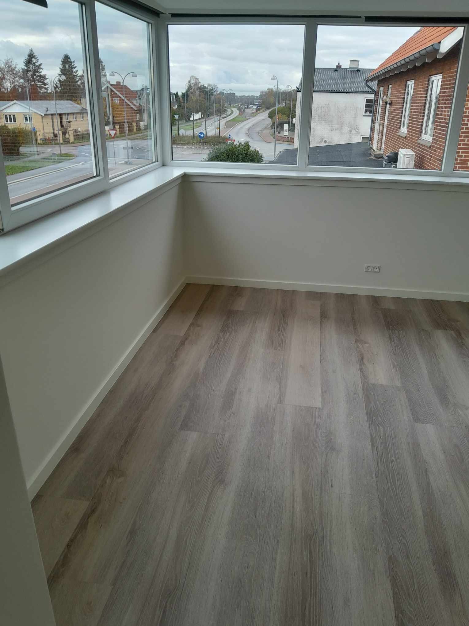 Jasnoszare panele podłogowe ułożone w pokoju z dużym oknem, widok na ulicę i domy z zewnątrz, ściany pomalowane na biało, widoczne gniazdka elektryczne przy podłodze.