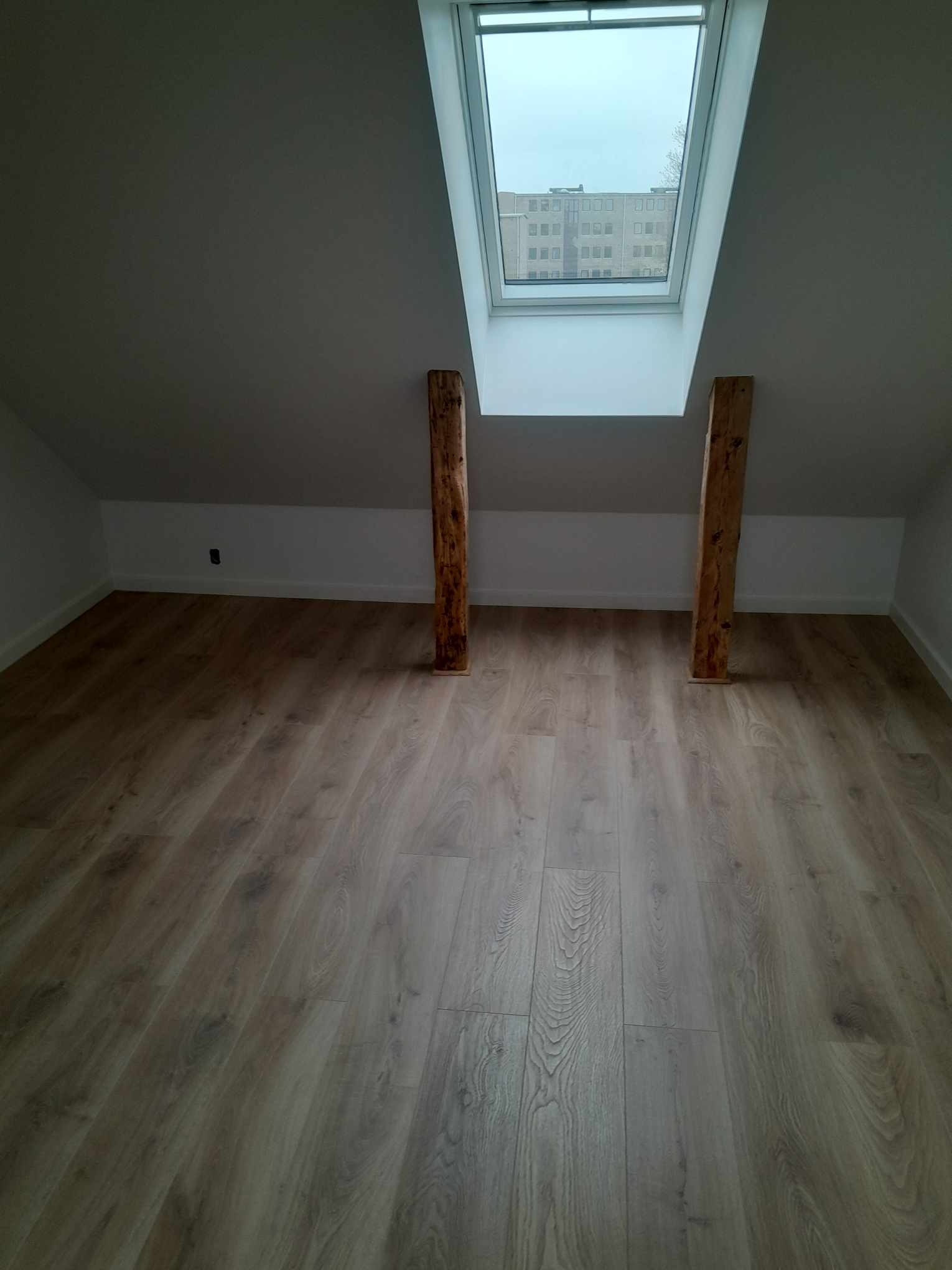 Poddasze z nowo ułożonymi panelami podłogowymi w odcieniach szarości, oświetlone przez okno dachowe z widokiem na budynek mieszkalny, wsparte dwoma drewnianymi słupami.