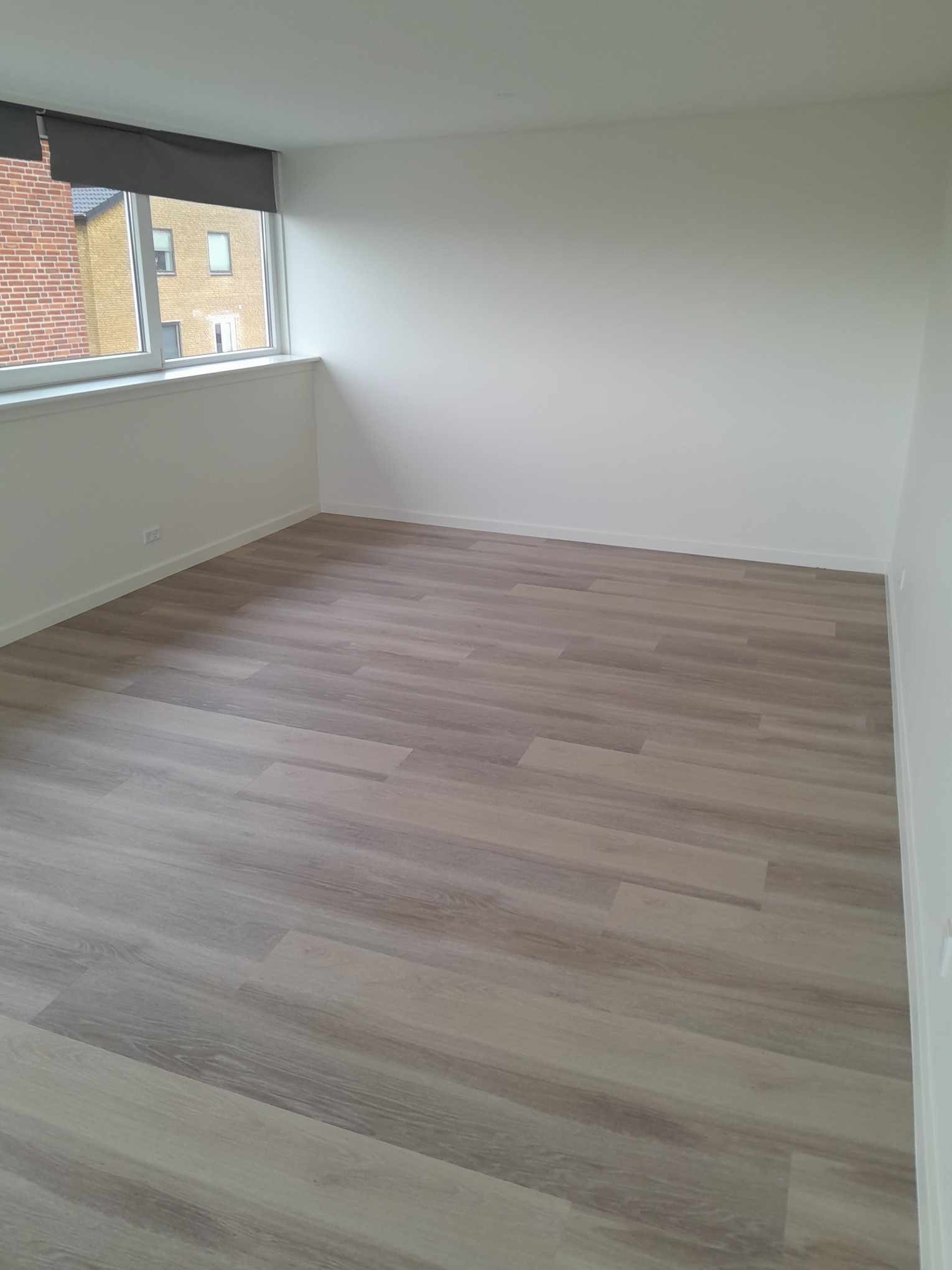Pusty pokój z nowo ułożonymi panelami podłogowymi w odcieniach szarości i beżu, białe ściany, okno z roletą w górnej części kadru.