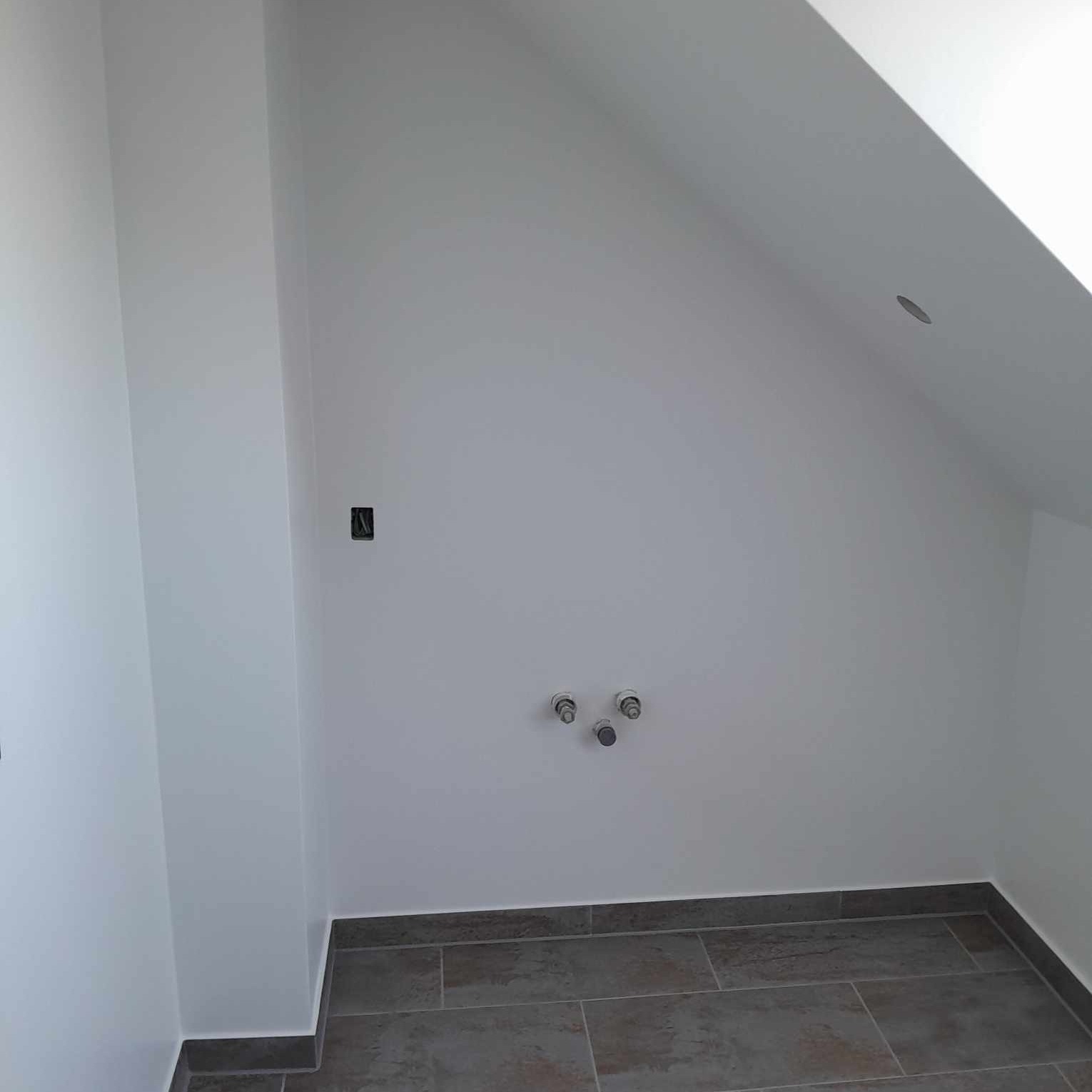 Świeżo pomalowane, białe poddasze ze skośnym sufitem, szare płytki podłogowe i wystające rury instalacji wodnej, widoczny mały otwór na gniazdko.