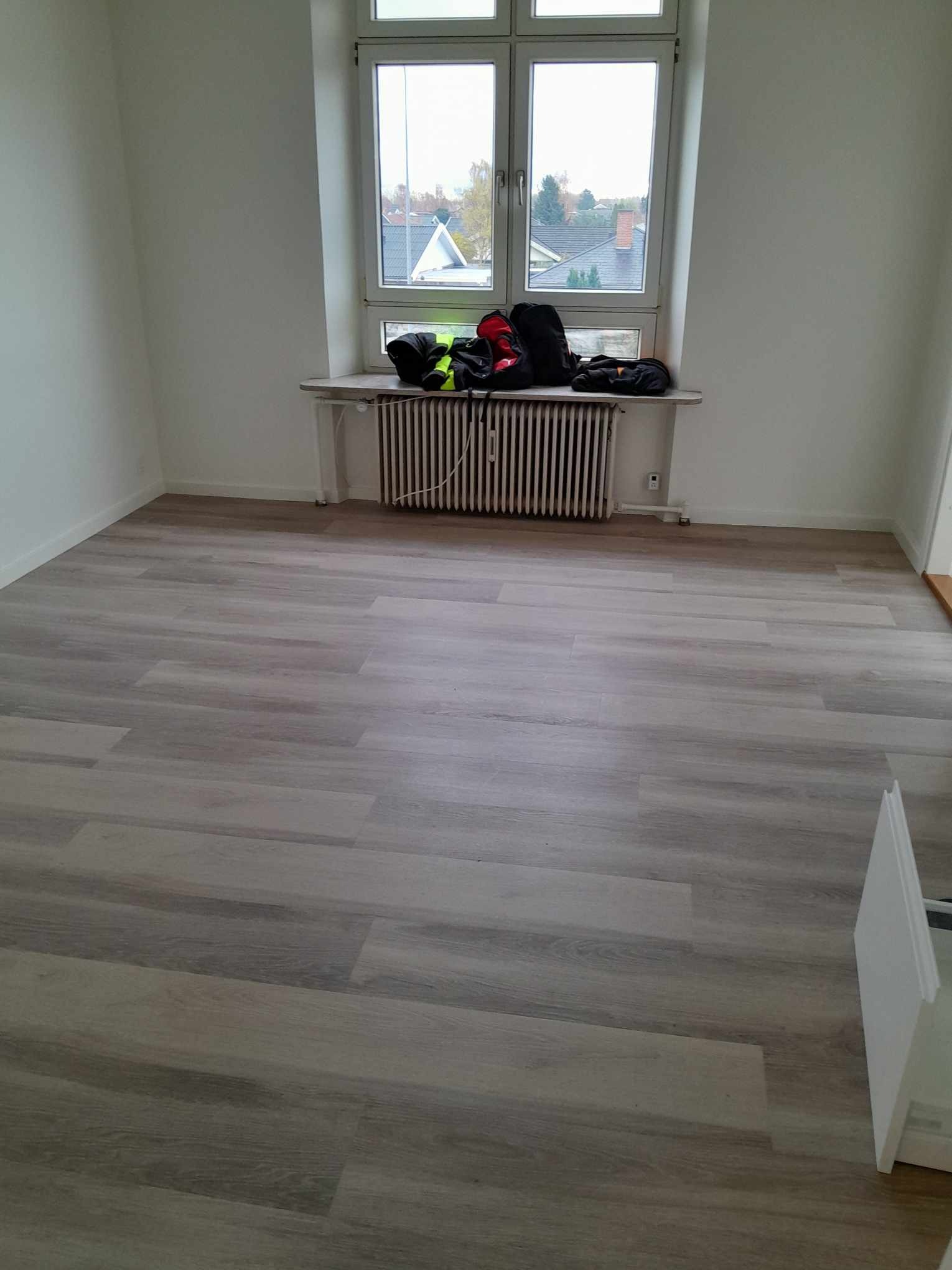 Jasnoszara podłoga z paneli ułożona w pustym pokoju z białymi ścianami, grzejnikiem pod oknem i torbami na parapecie.