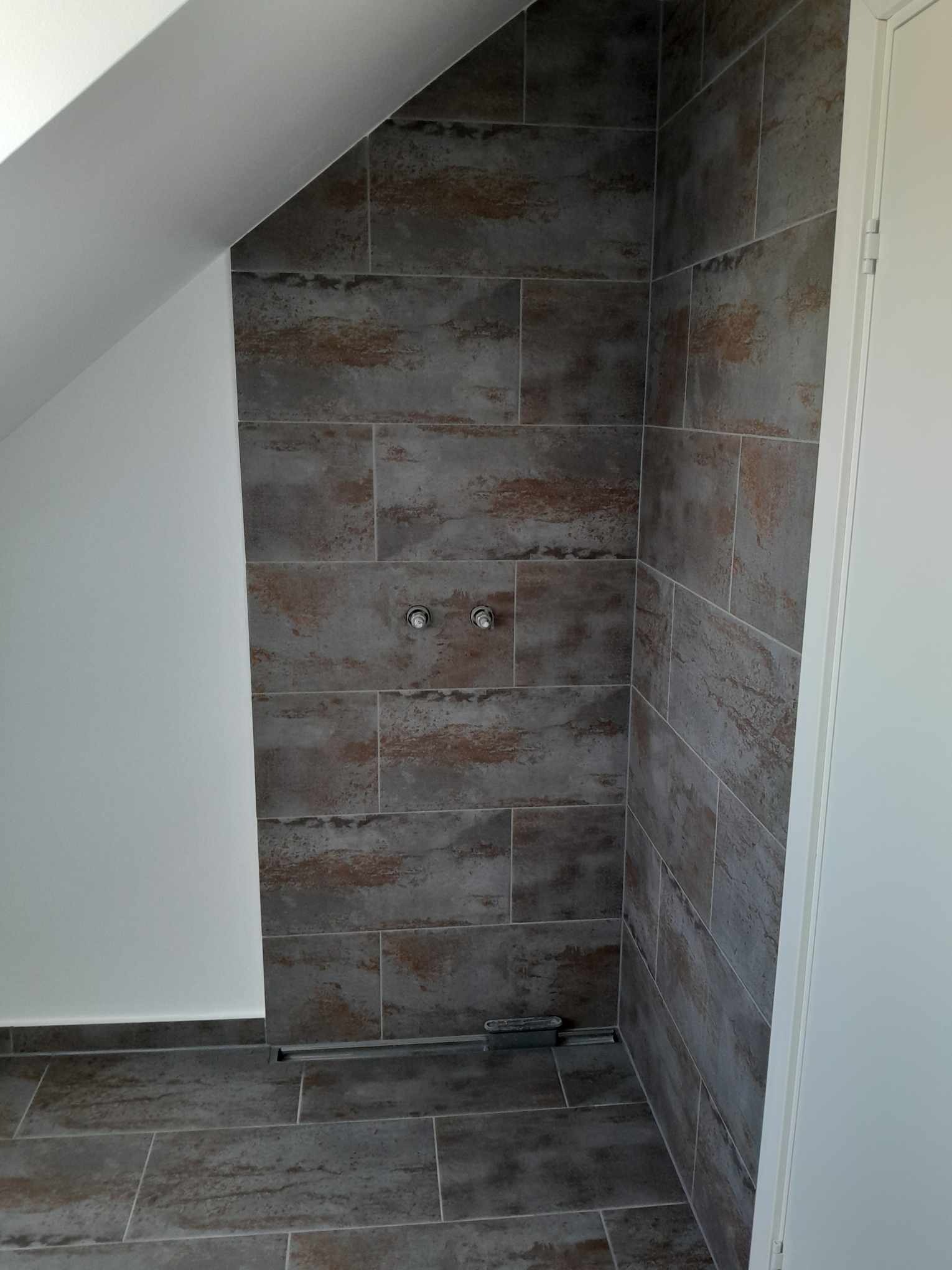 Ukończona kabina prysznicowa w łazience na poddaszu, wyłożona szarymi płytkami imitującymi kamień z brązowymi akcentami, widoczny odpływ liniowy i przygotowane przyłącza wodne.
