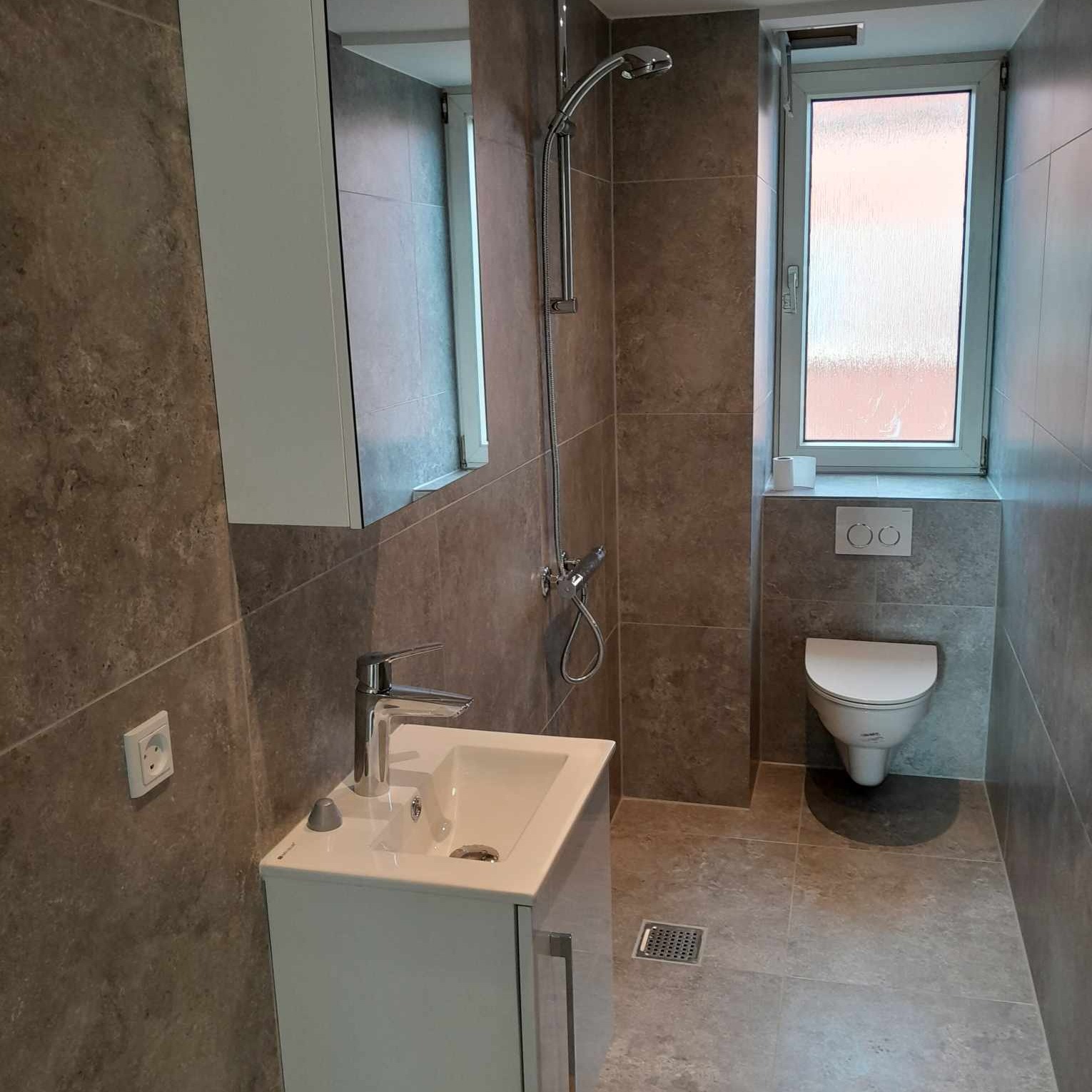 Nowoczesna łazienka z szarymi płytkami, wiszącą toaletą, kabiną prysznicową bez brodzika i szafką z umywalką, oświetlona światłem dziennym z okna.