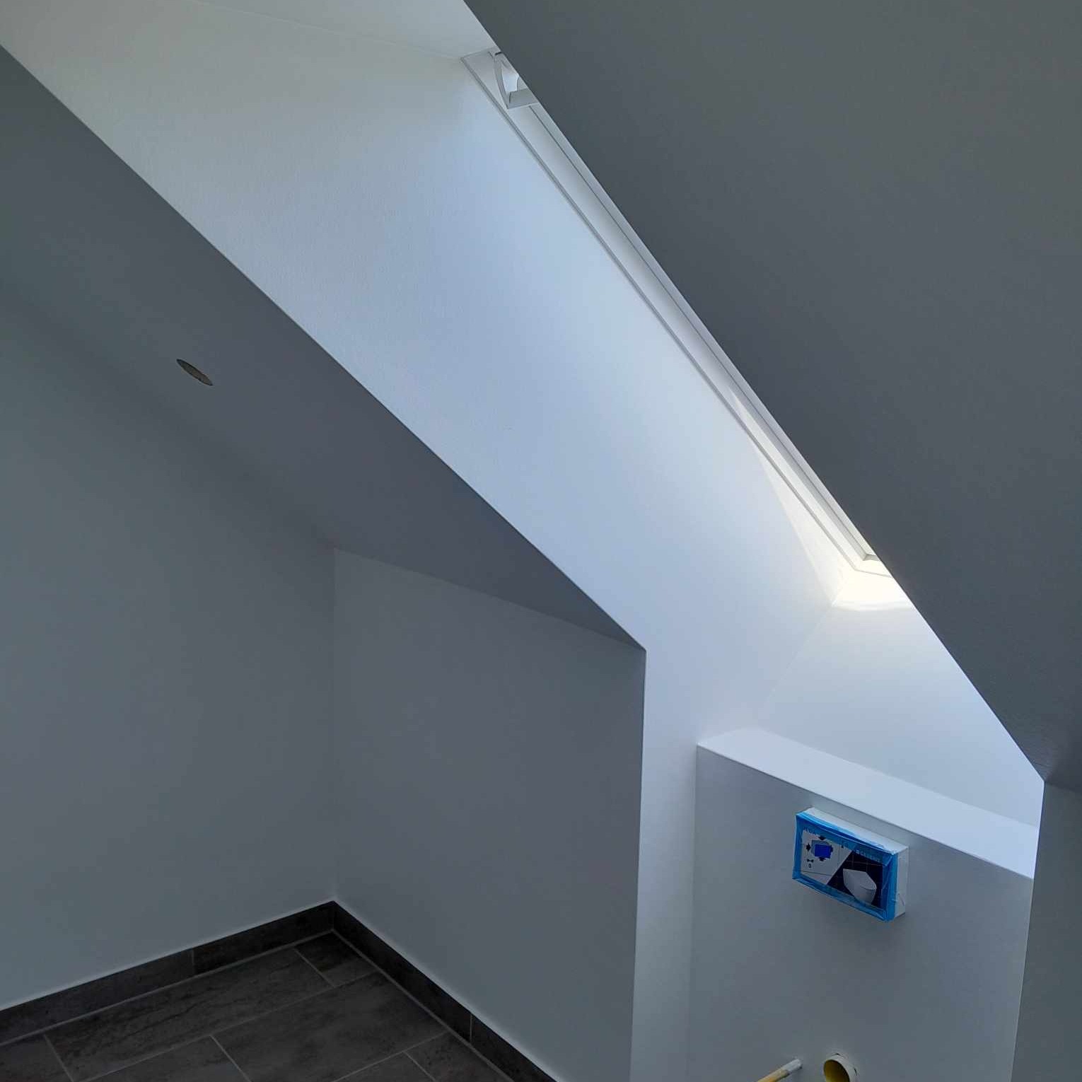 Poddasze po odświeżeniu: białe ściany i sufit, okno dachowe w skosie, szare płytki podłogowe, częściowo zamaskowana instalacja sanitarna na ścianie.