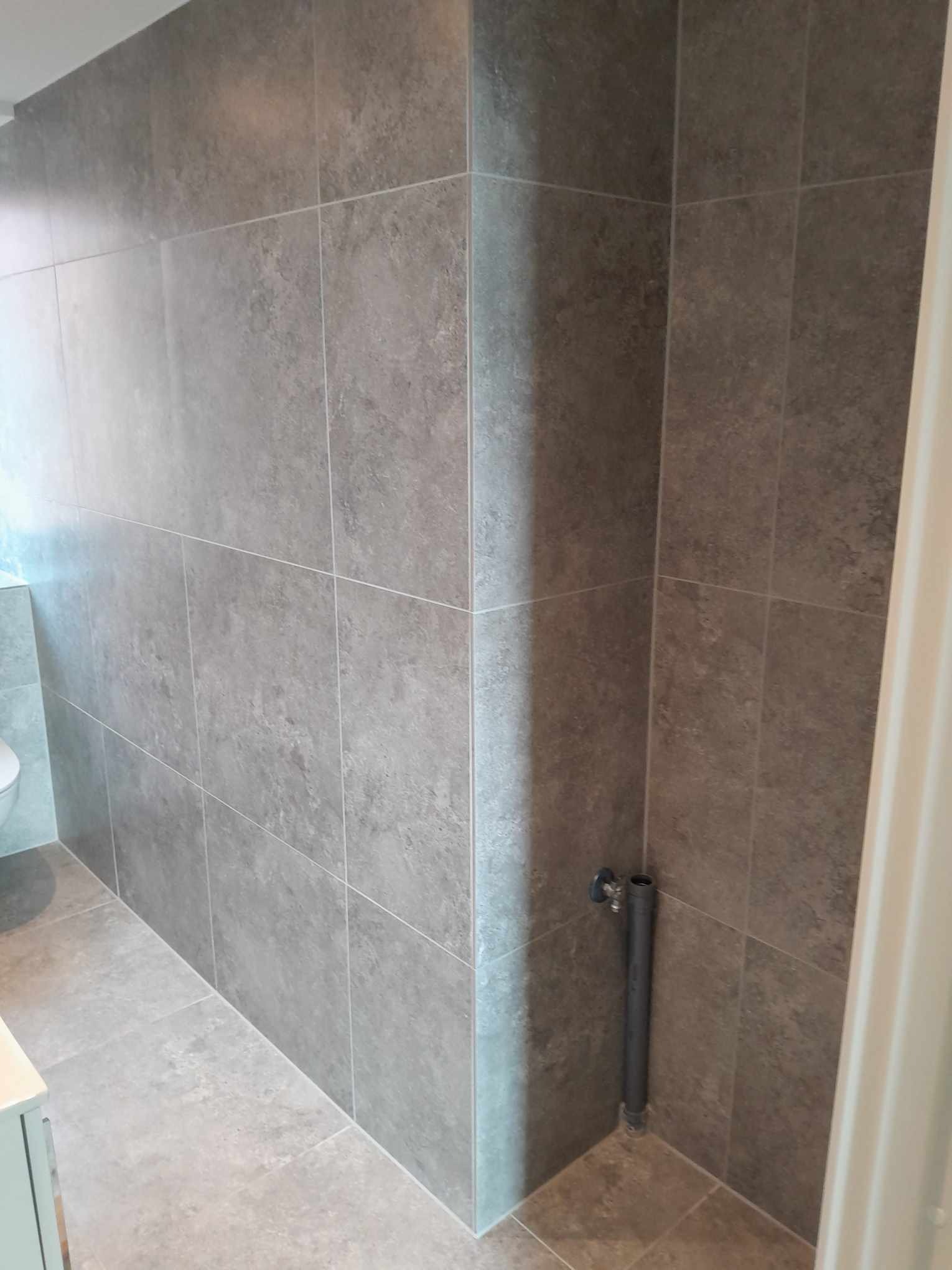 Szare, matowe płytki ceramiczne wielkoformatowe na ścianach i podłodze w rogu łazienki, widoczna rura instalacyjna przy ścianie.