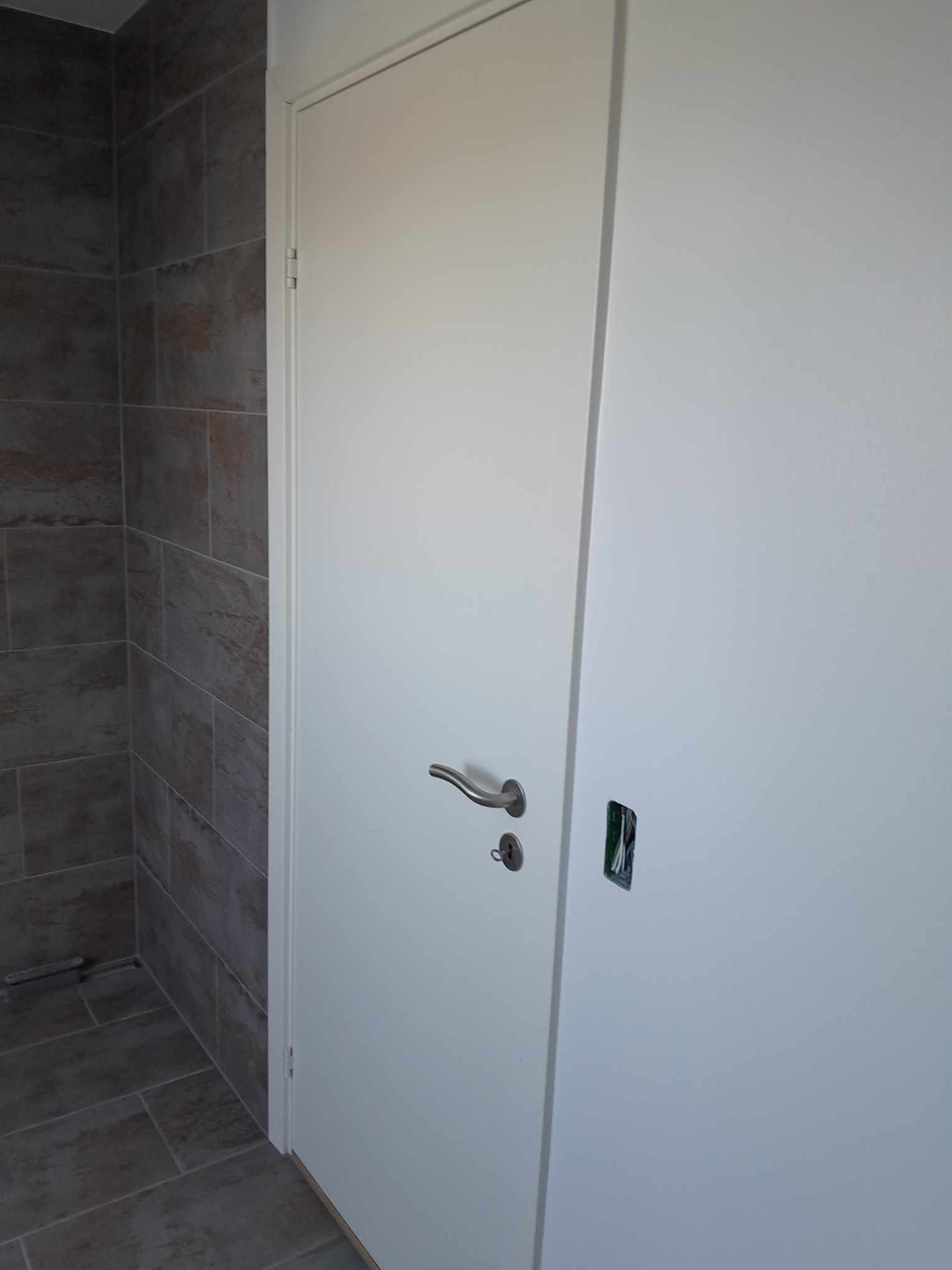 Białe drzwi z chromowaną klamką, zamontowane w łazience wykończonej szarymi, matowymi płytkami imitującymi kamień; widoczny otwór na zamek elektroniczny z wystającymi przewodami.