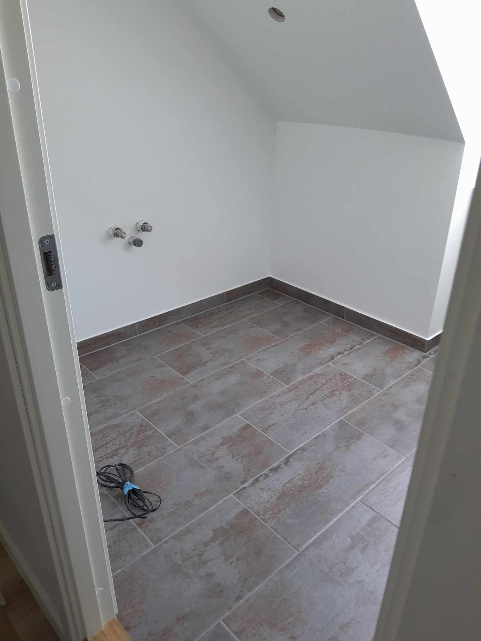 Płytki podłogowe w odcieniach szarości i brązu w małym pomieszczeniu, widoczne przyłącza wodne na ścianie, kabel elektryczny leżący na podłodze, widok zza uchylonych drzwi.
