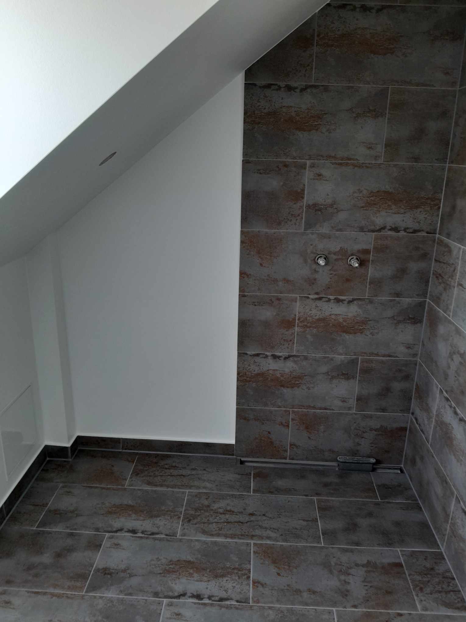 Wykończona łazienka na poddaszu z szarymi płytkami imitującymi kamień na podłodze i ścianach, widoczne dwa chromowane przyłącza wodne i odpływ liniowy w podłodze.