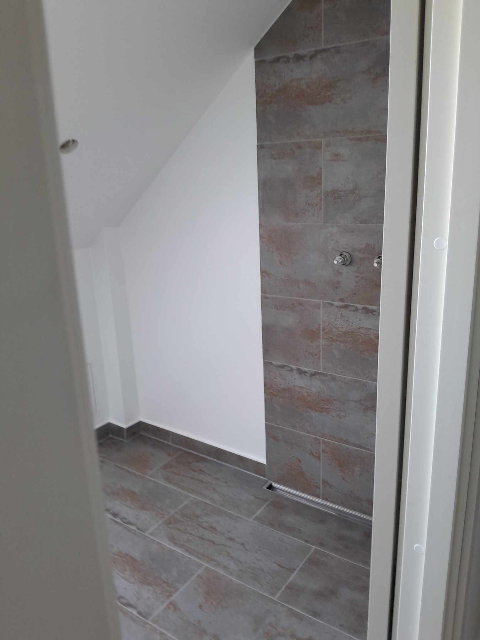 Wnętrze łazienki w trakcie wykańczania, widoczne szare płytki na ścianie i podłodze, białe ściany, zamontowany odpływ liniowy, oraz fragment ramy drzwi.