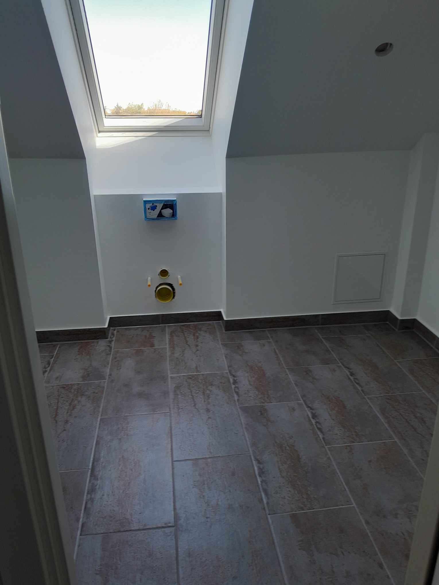Surowa łazienka z szarymi płytkami podłogowymi, przygotowana pod montaż toalety podtynkowej, widoczne rury kanalizacyjne i okno dachowe.