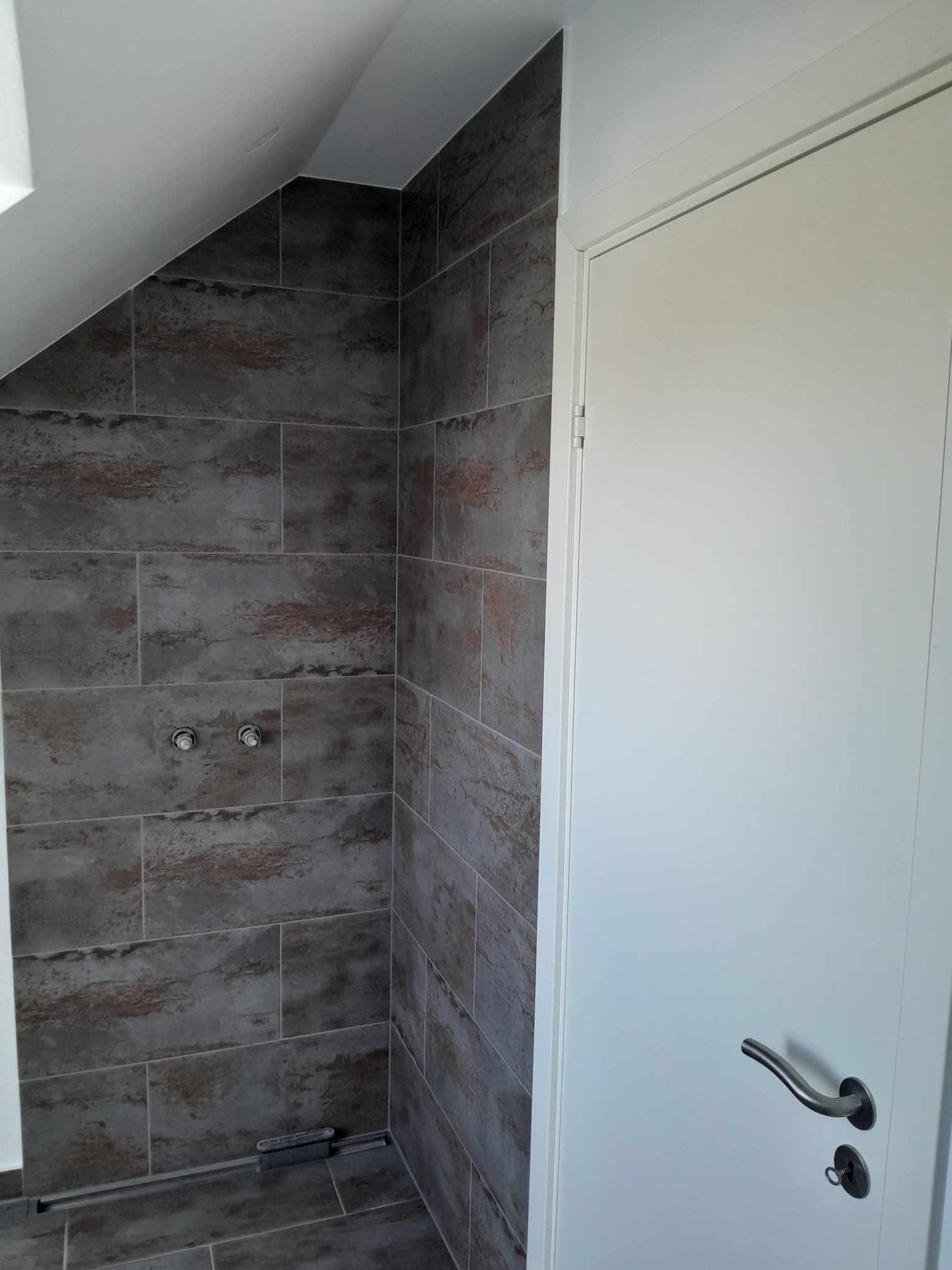 Wykończona kabina prysznicowa z ciemnoszarymi płytkami imitującymi kamień, widoczne chromowane zawory i odpływ liniowy, obok białe drzwi wejściowe do łazienki z srebrną klamką.