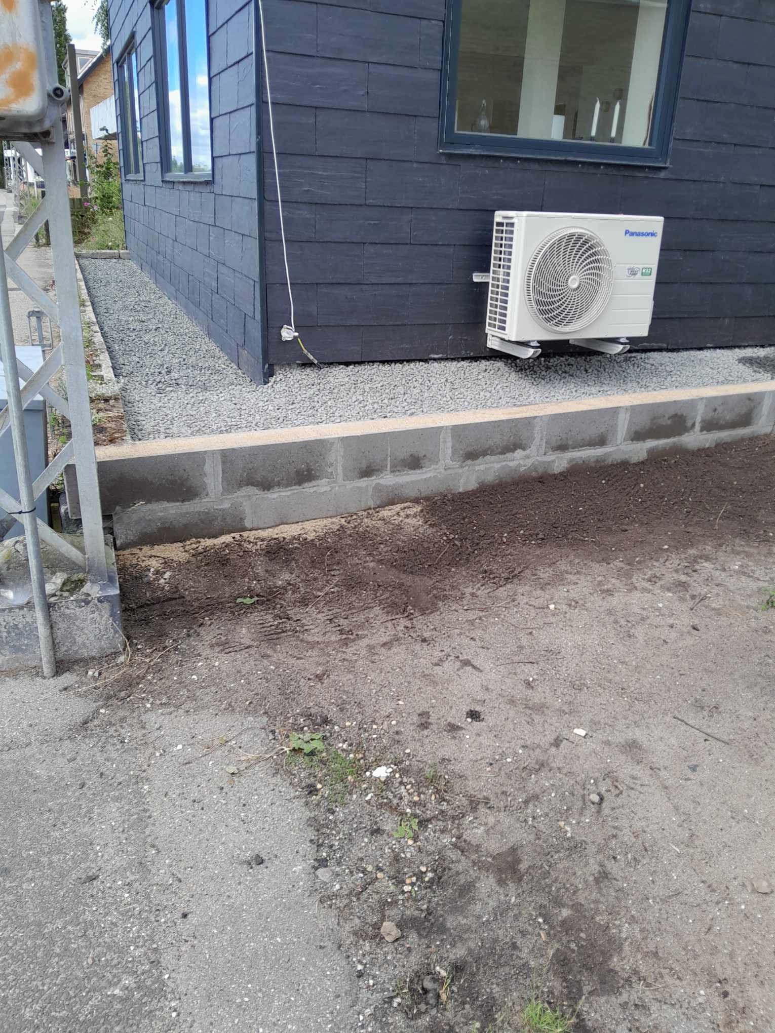 Ciemna elewacja domu z zamontowanym klimatyzatorem Panasonic, obok widoczny fundament z betonowych bloczków i nasypany żwir.