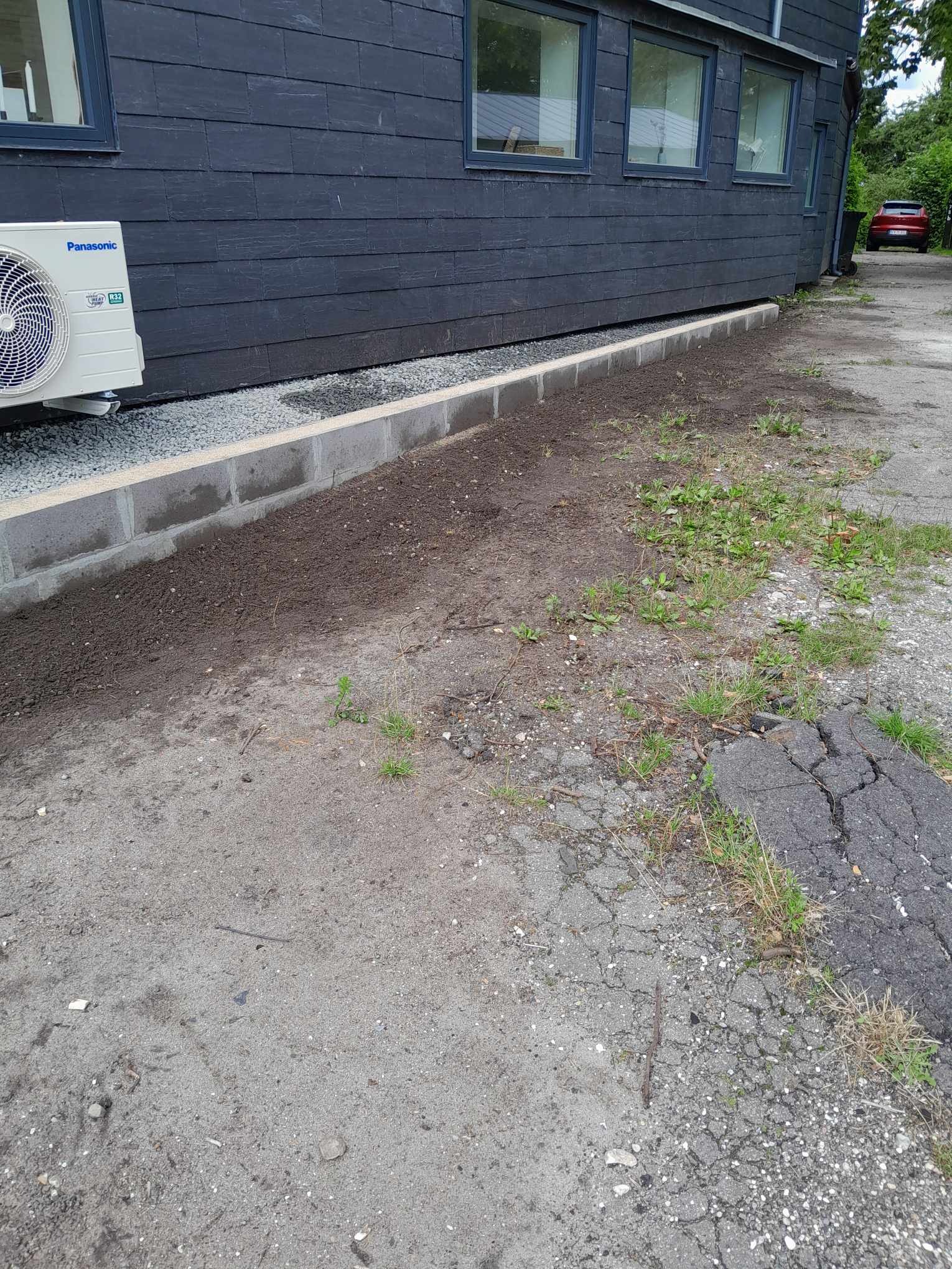 Przygotowanie terenu pod trawnik przy ciemnej elewacji domu, widoczny klimatyzator Panasonic i częściowo ułożony obrzeże z betonowych bloczków.