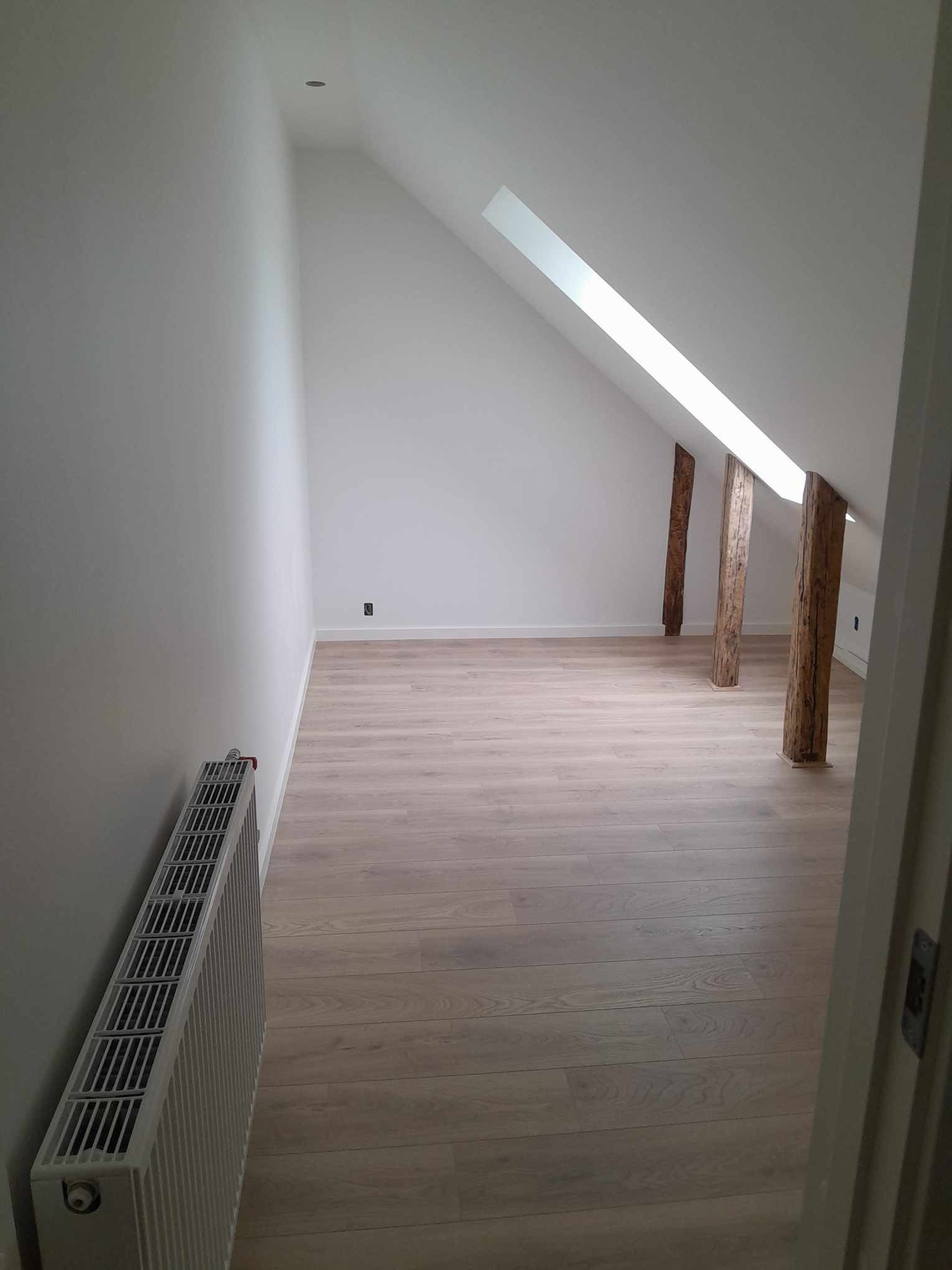 Poddasze po remoncie w Sławnie: świeżo pomalowane ściany na biało, jasne panele podłogowe, odsłonięte drewniane belki konstrukcyjne i grzejnik przy ścianie.