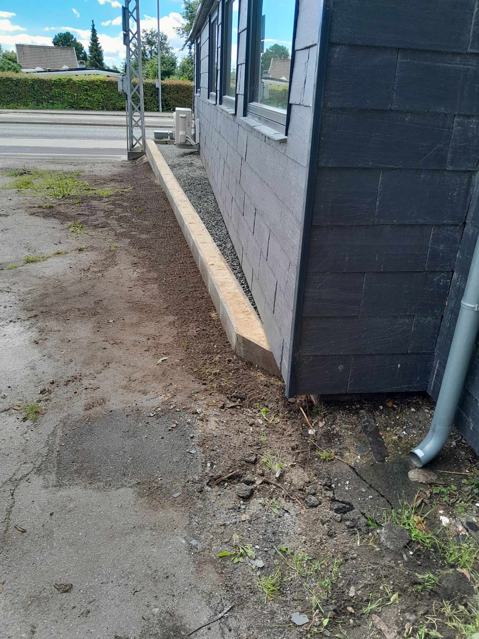 Elewacja budynku wykończona ciemnoszarym łupkiem, z widoczną betonową obwódką i rynną spustową, otoczona ziemią i fragmentem chodnika.