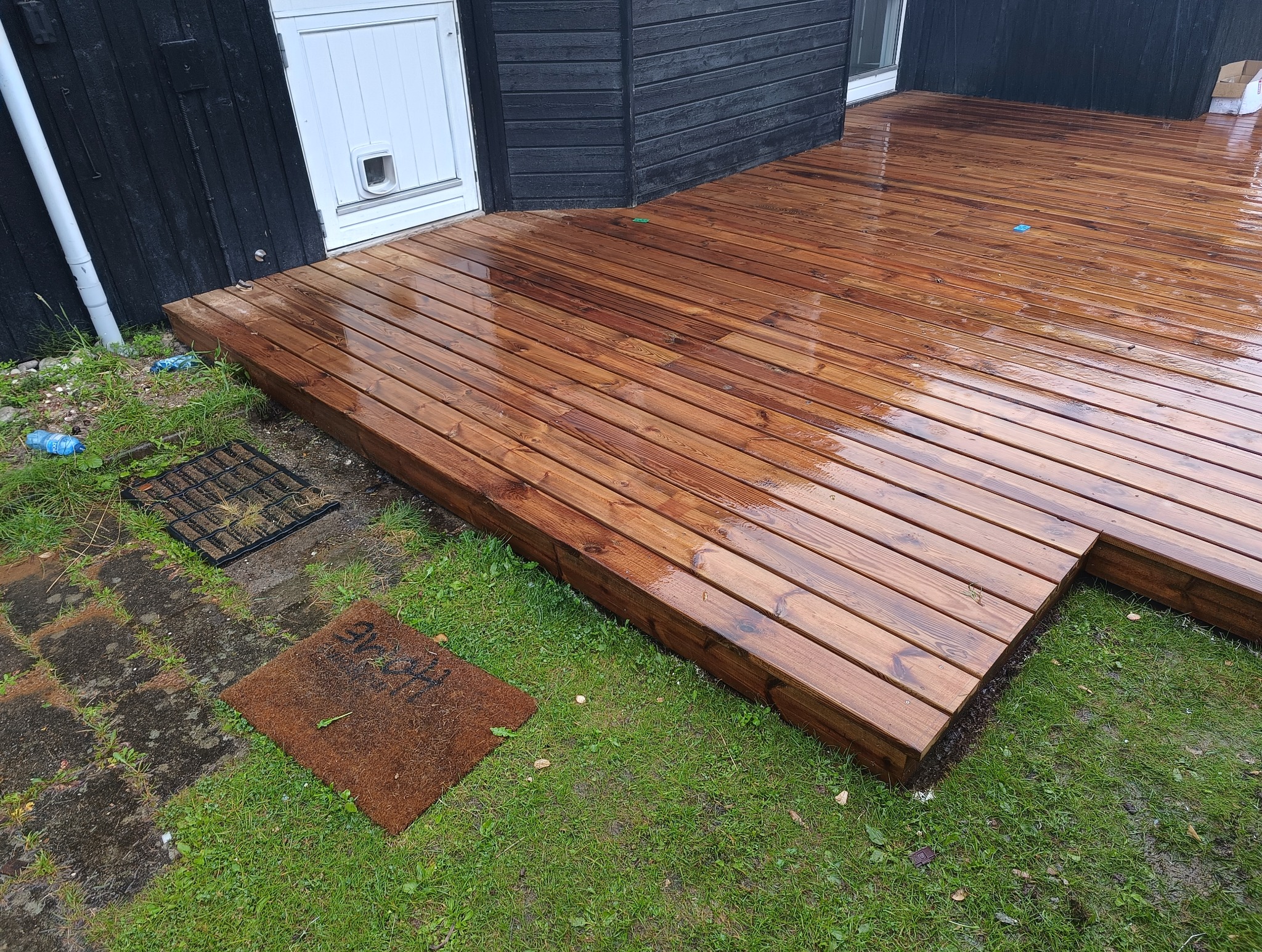 Świeżo zaimpregnowany, mokry taras drewniany z desek, widok z góry na fragment ogrodu i czarną elewację budynku z białymi drzwiami.