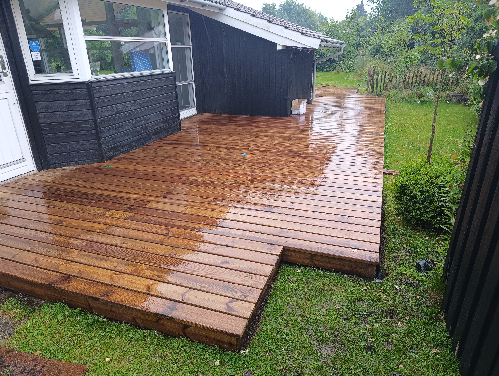 Świeżo zaimpregnowany, drewniany taras przylegający do czarnego budynku z białymi ramami okiennymi, otoczony zielonym trawnikiem i roślinnością.