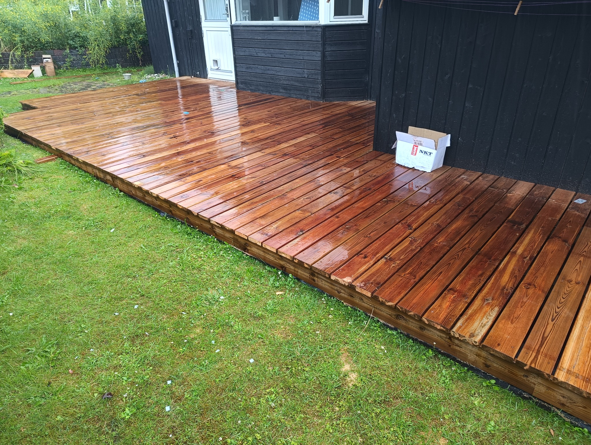 Świeżo zaimpregnowany, mokry drewniany taras przylegający do czarnej ściany budynku, obok kartonowe pudełko z napisem NKT.