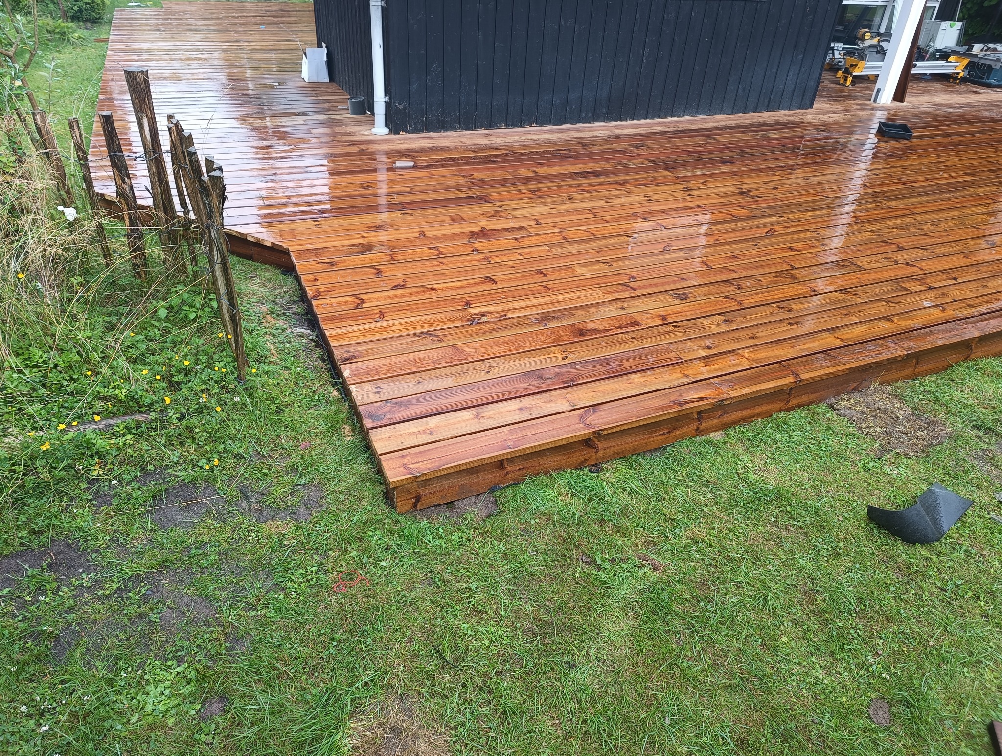 Świeżo polakierowany drewniany taras przylegający do czarnej ściany budynku, z widoczną zieloną trawą i prowizorycznym płotkiem z gałęzi.