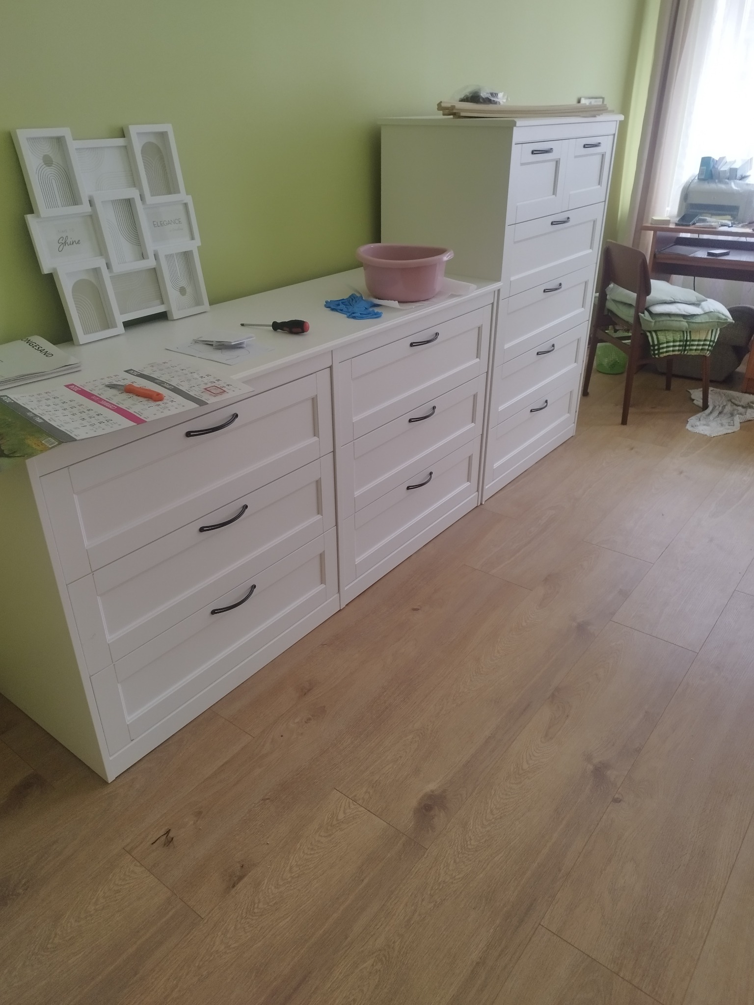 Białe komody z szufladami na drewnianej podłodze, na blacie leżą narzędzia, kalendarz, rękawice robocze i miska, na ścianie dekoracyjne ramki.