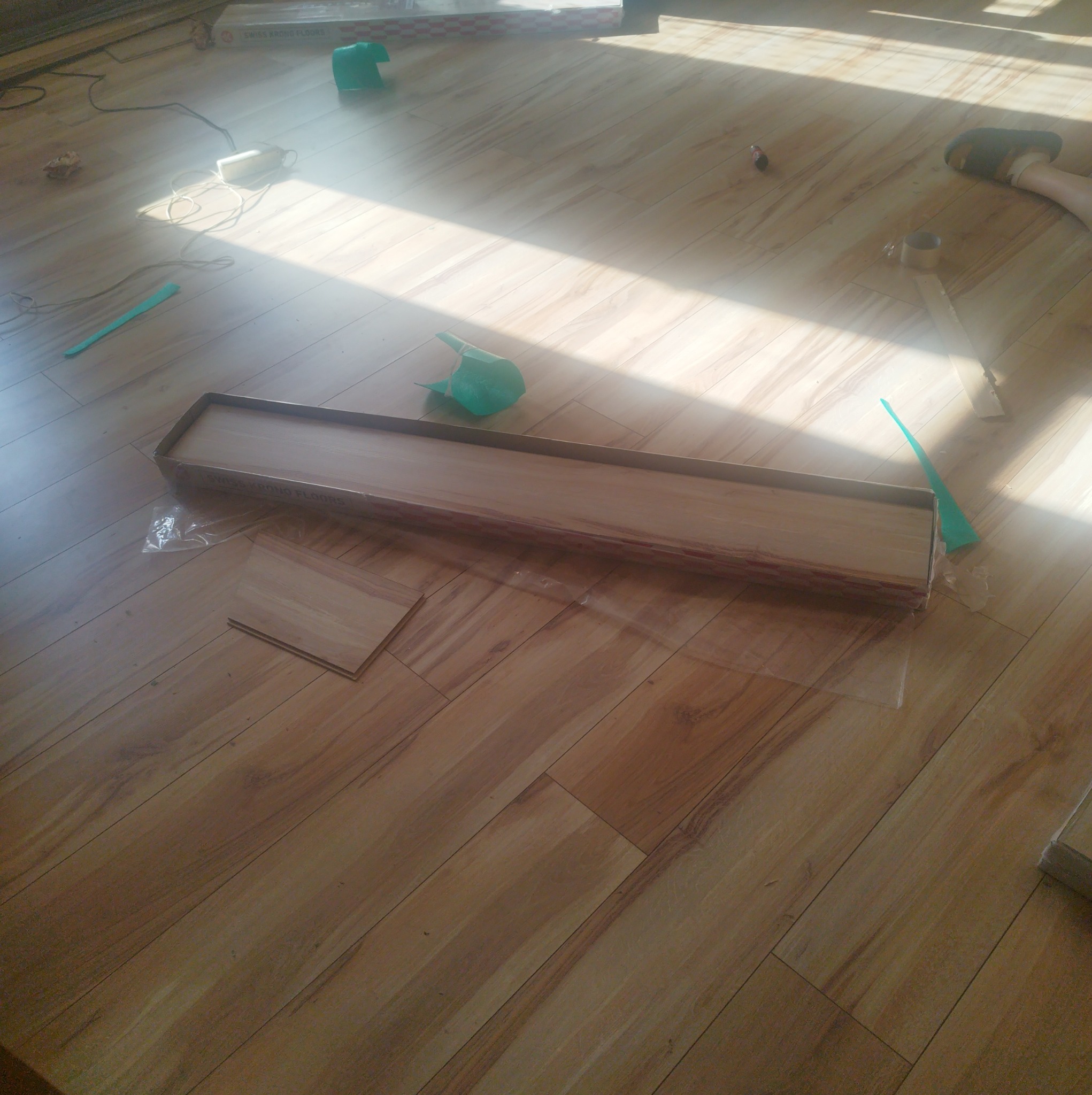 Rozpakowane panele podłogowe w kartonie, widoczne akcesoria montażowe i fragment stopy w klapku na nowej podłodze.